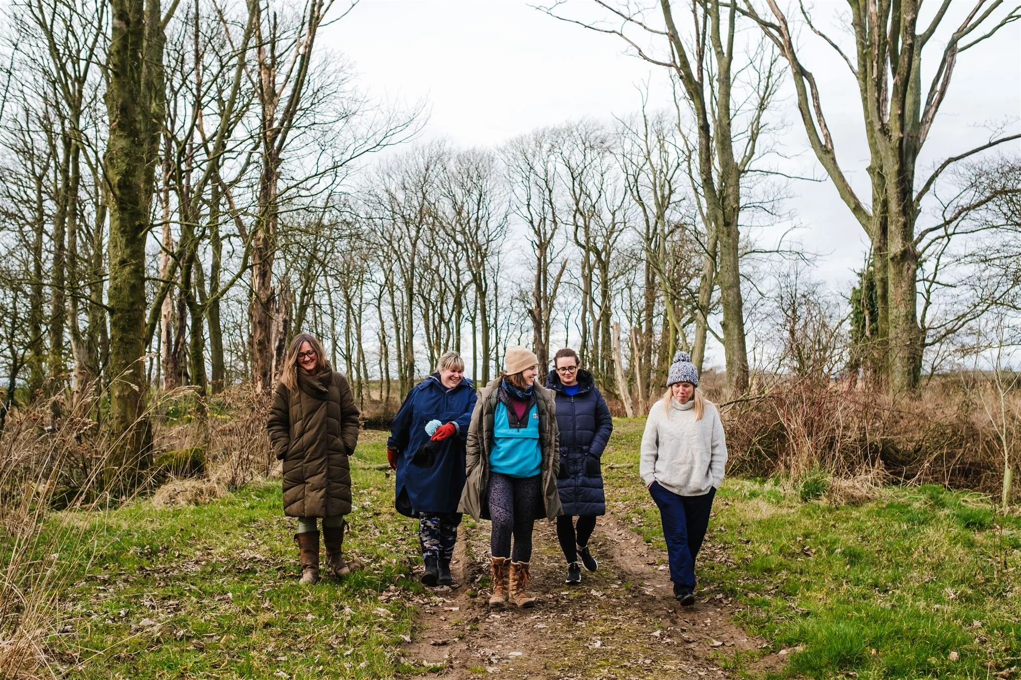 Abi Adams and four other women are in a forest walking. They are all smiling, laughing, and looking away from the camera.