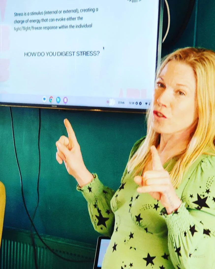 Abi Adams is speaking and gesturing with her hands whilst standing in front of a mounted screen with text on it.