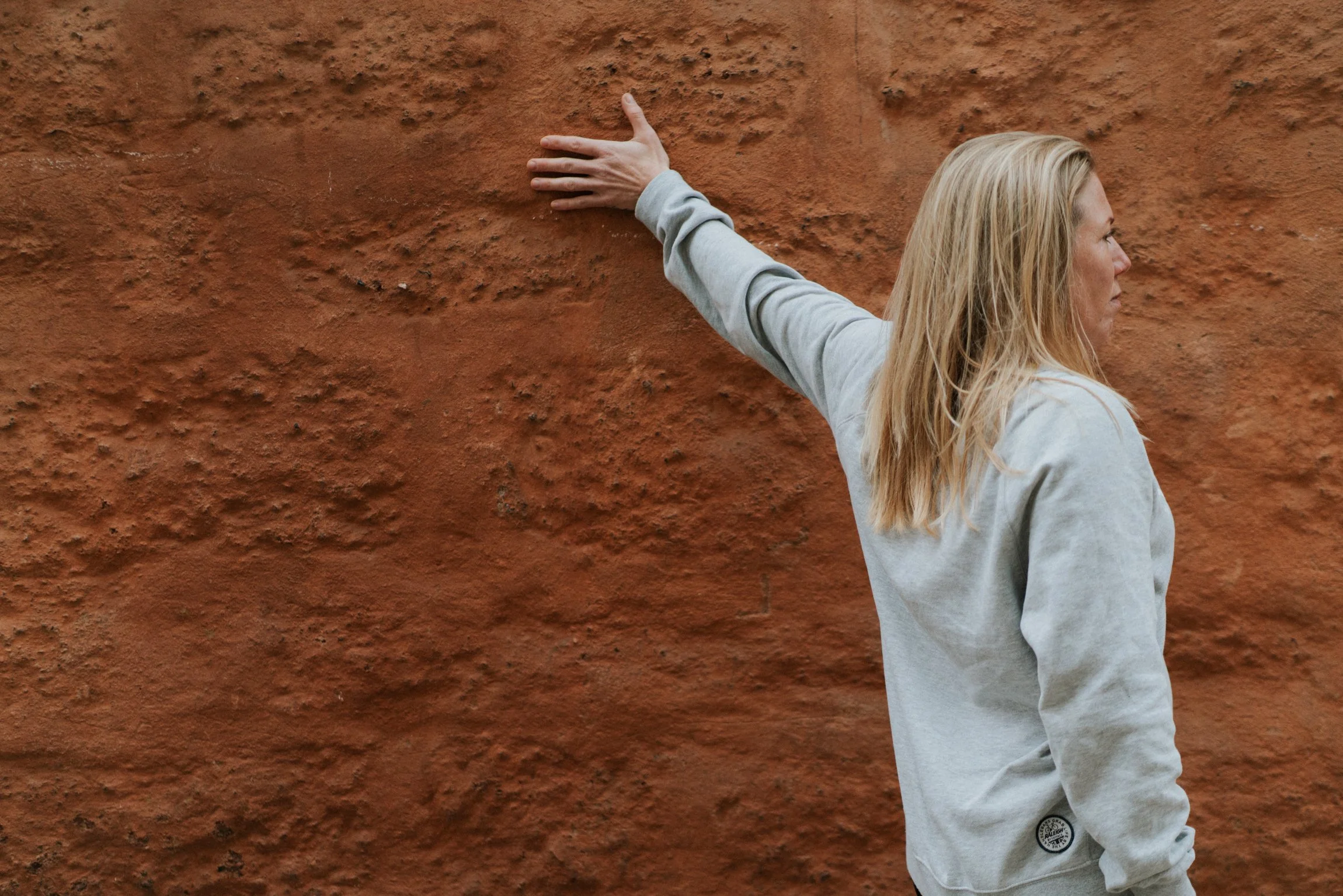 Abi Adams is standing in front of a terracotta wall, her left hand is on the wall and she is facing away from the wall and her left hand.
