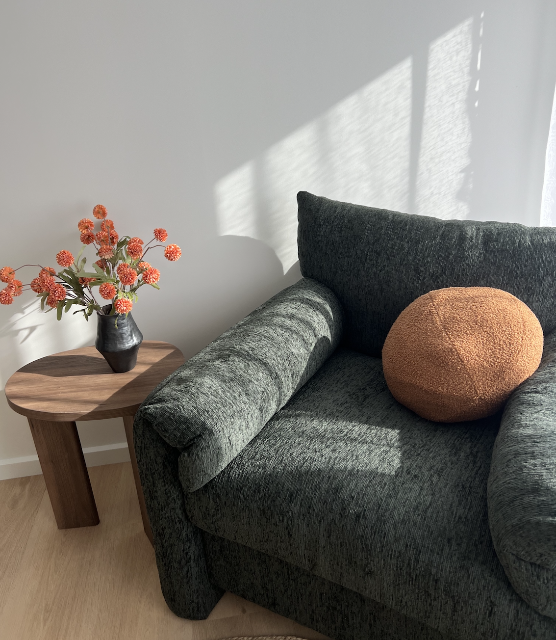 Therapy space with dark green upholstered sofa, an orange textured round pillow, a small wooden side table with a black vase holding orange flowers, and sunlight casting shadows on the wall.