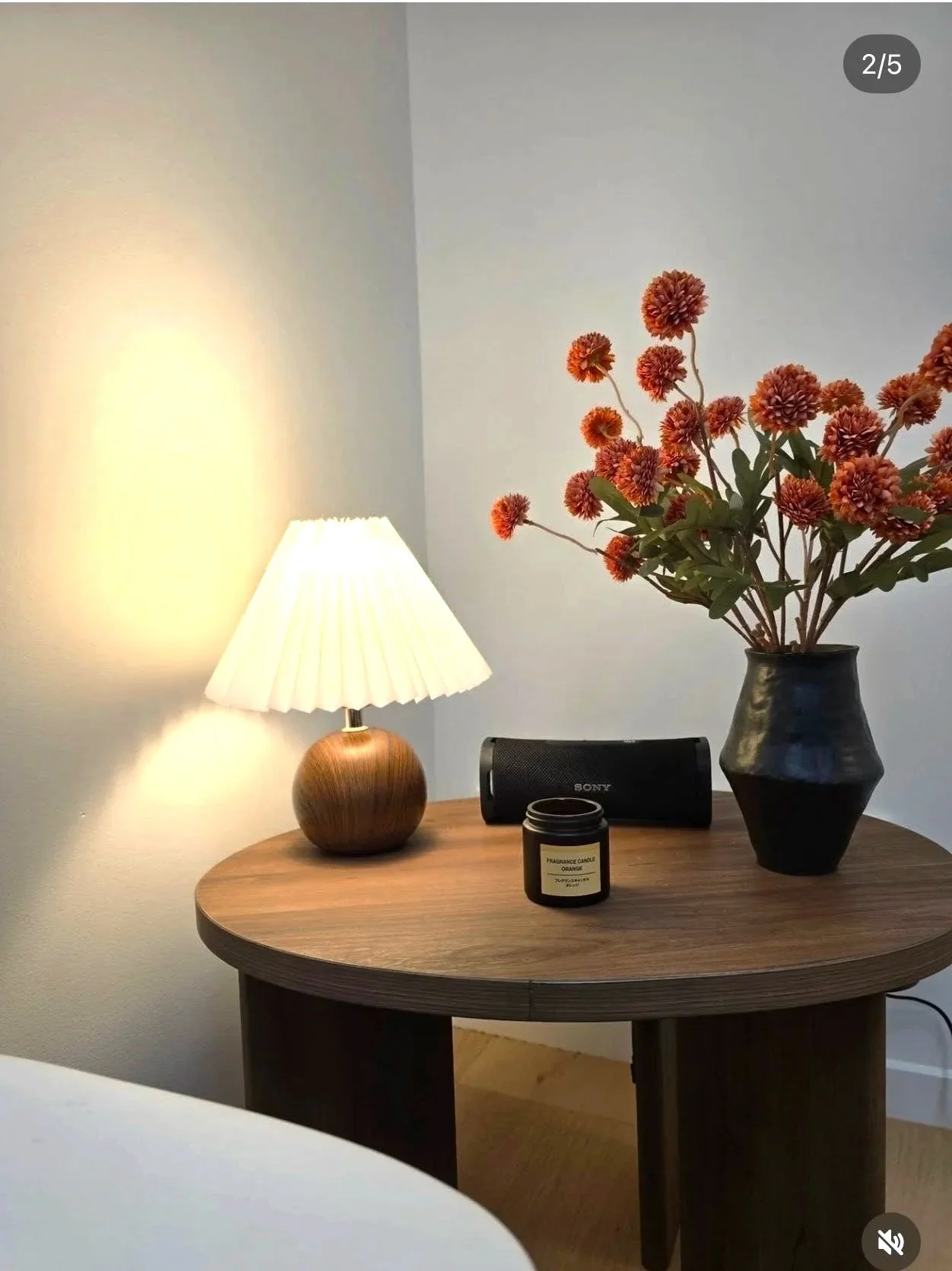 A wooden round side table with a lit table lamp, a black scented candle, a black portable speaker, and a black vase with pinkish-orange flowers.