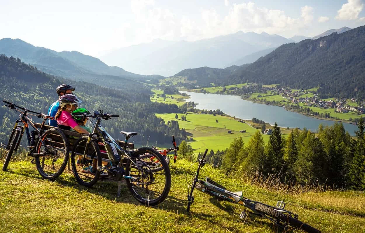 Two children with helmets sitting on mountain bikes and a mountain bike lying on the grass, overlooking a green valley with a lake, surrounded by forested mountains under a partly cloudy sky.