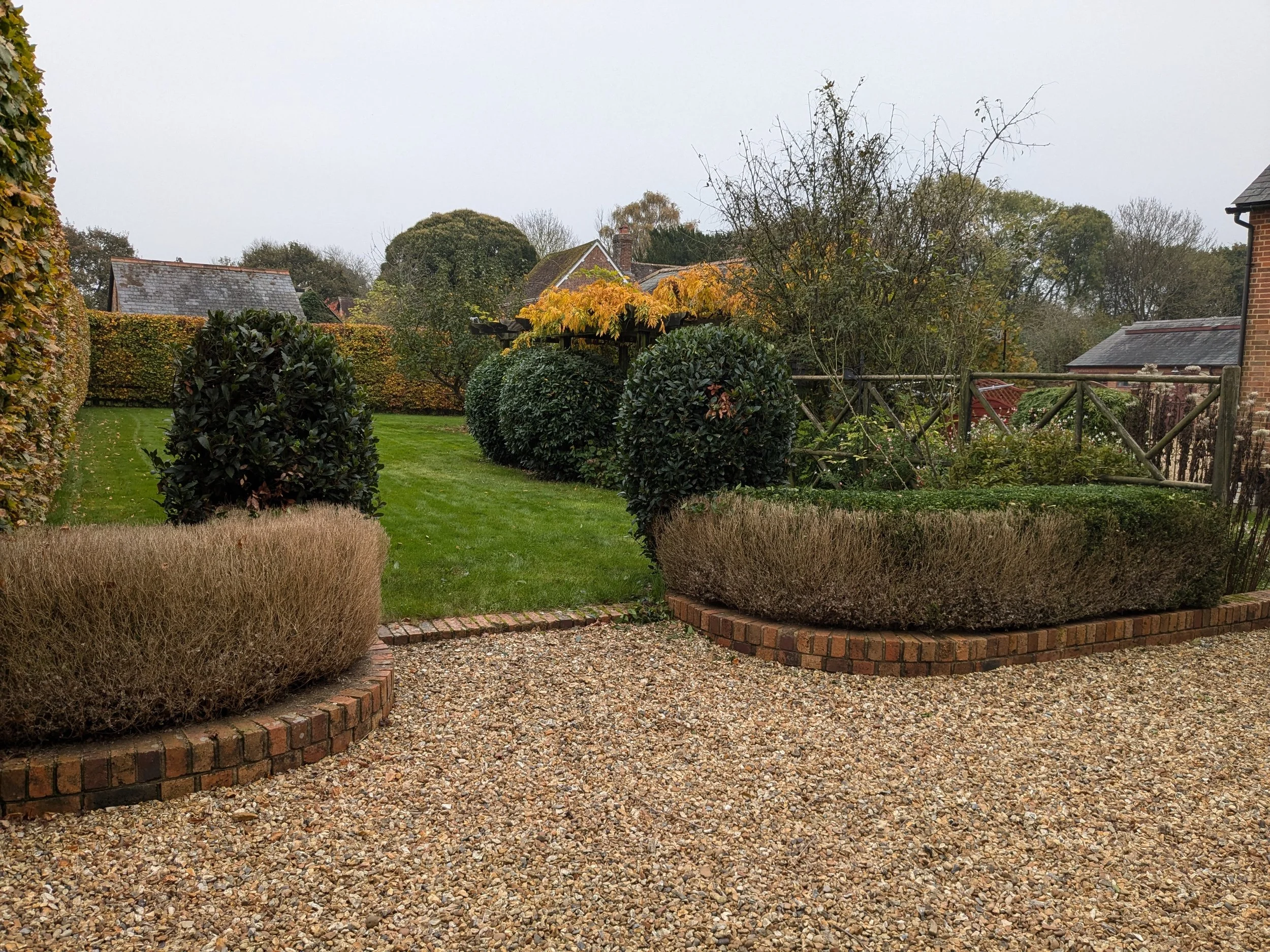 A garden with neatly trimmed bushes, a gravel pathway, and a grassy lawn with trees, shrubs, and a wooden fence in the background under an overcast sky.