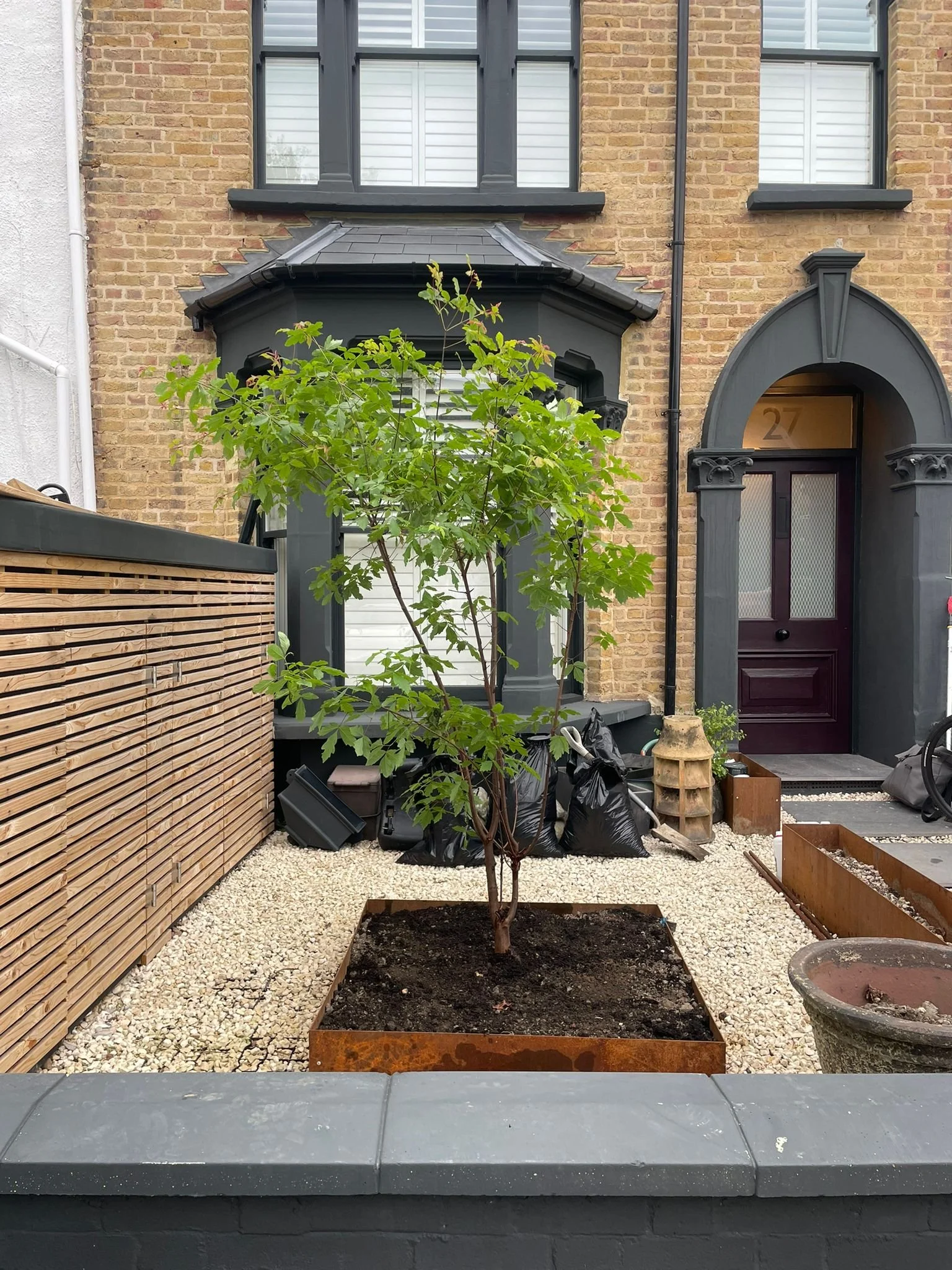A young tree planted in a square metal container in an urban yard. Surrounding gravel and some pots, with a brick building and black door in the background.