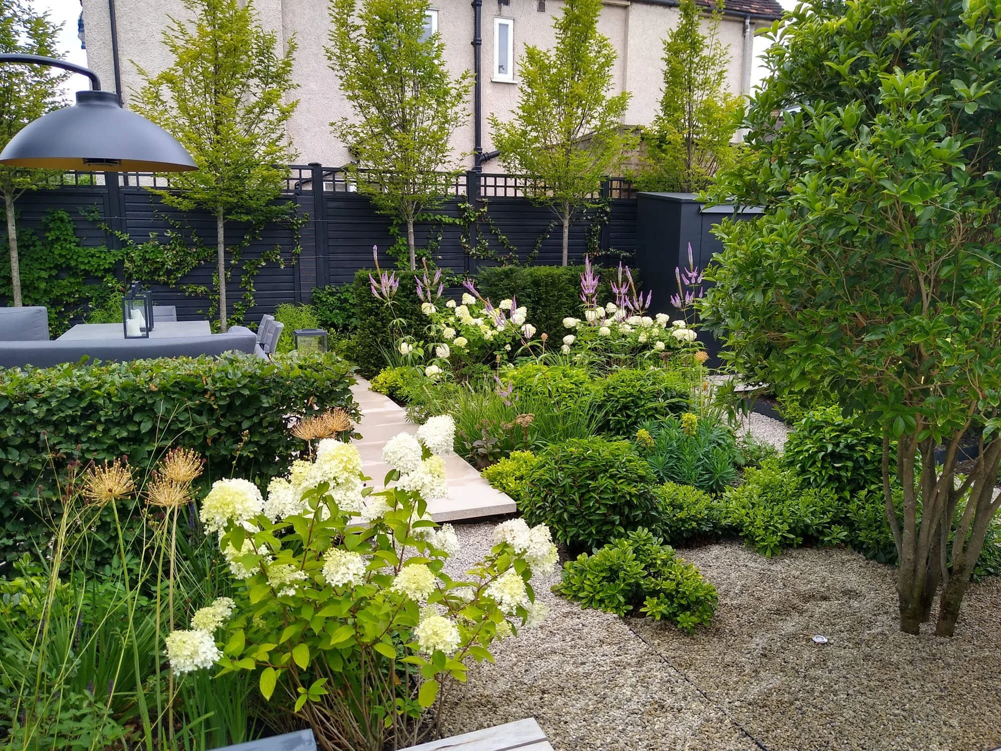 A well-maintained backyard garden with lush green bushes, flowering plants, and trees. There is a gravel path and a seating area with a table and chairs under a large black patio umbrella. The garden is enclosed by a black fence with small trees growing along it. The background includes a white house with windows.