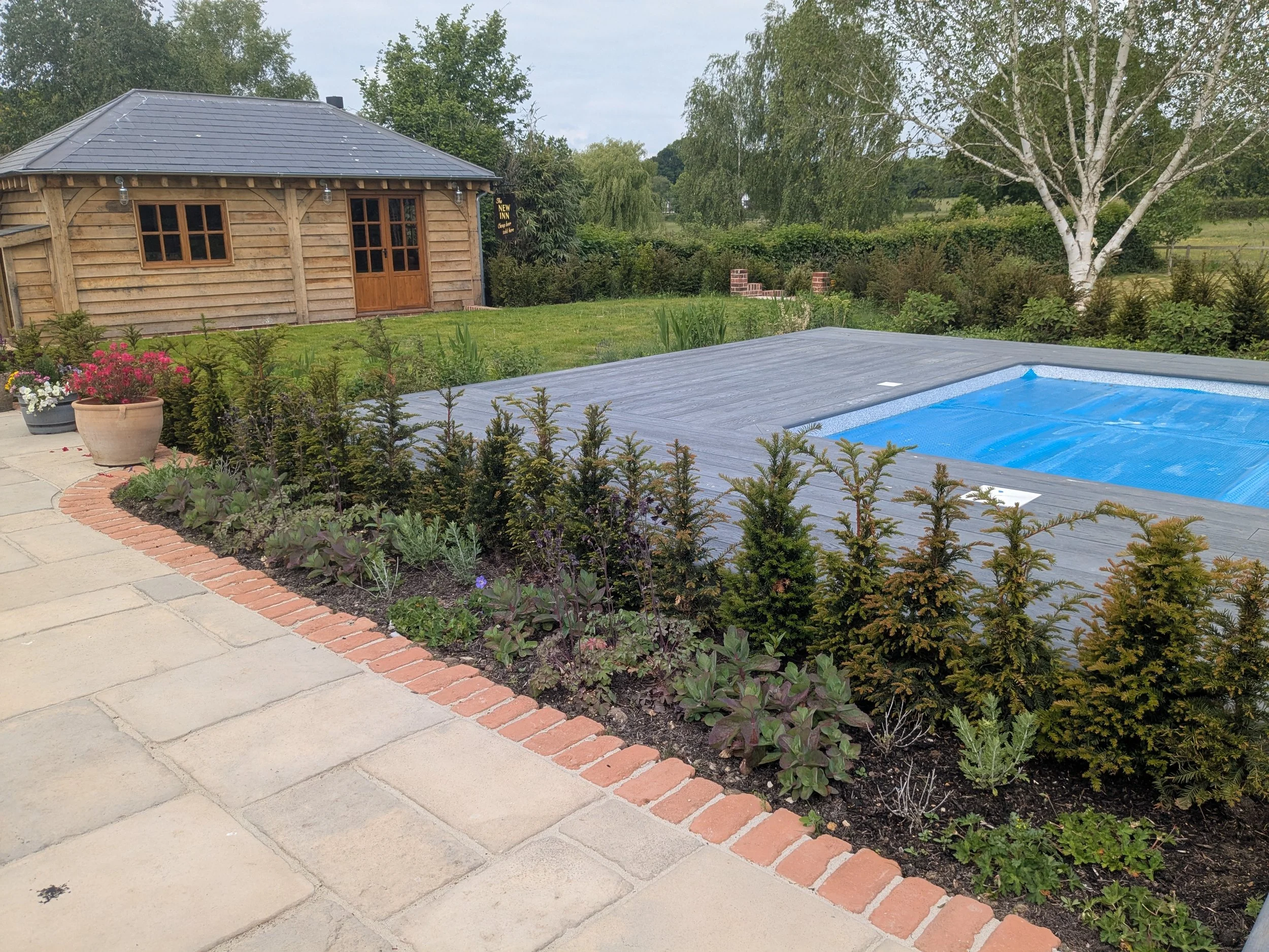 Backyard with stone patio, garden bed with shrubs and purple flowers, wooden shed, and a swimming pool covered with a blue tarp, surrounded by trees and greenery.