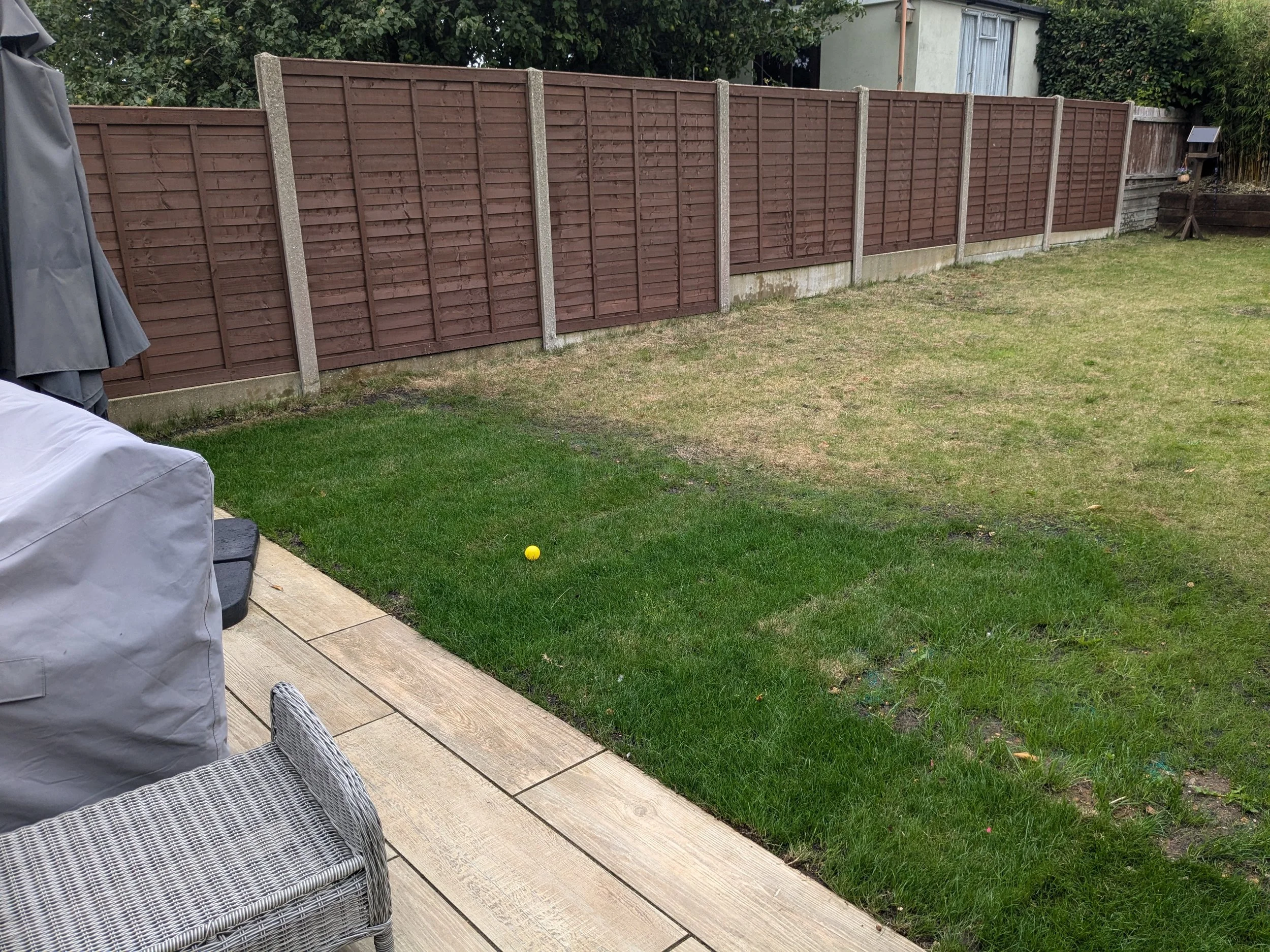 View of a backyard with a wooden deck, lawn, and a brown wooden fence. A yellow ball is on the grass, and a gray patio umbrella and outdoor furniture are partially visible on the deck.