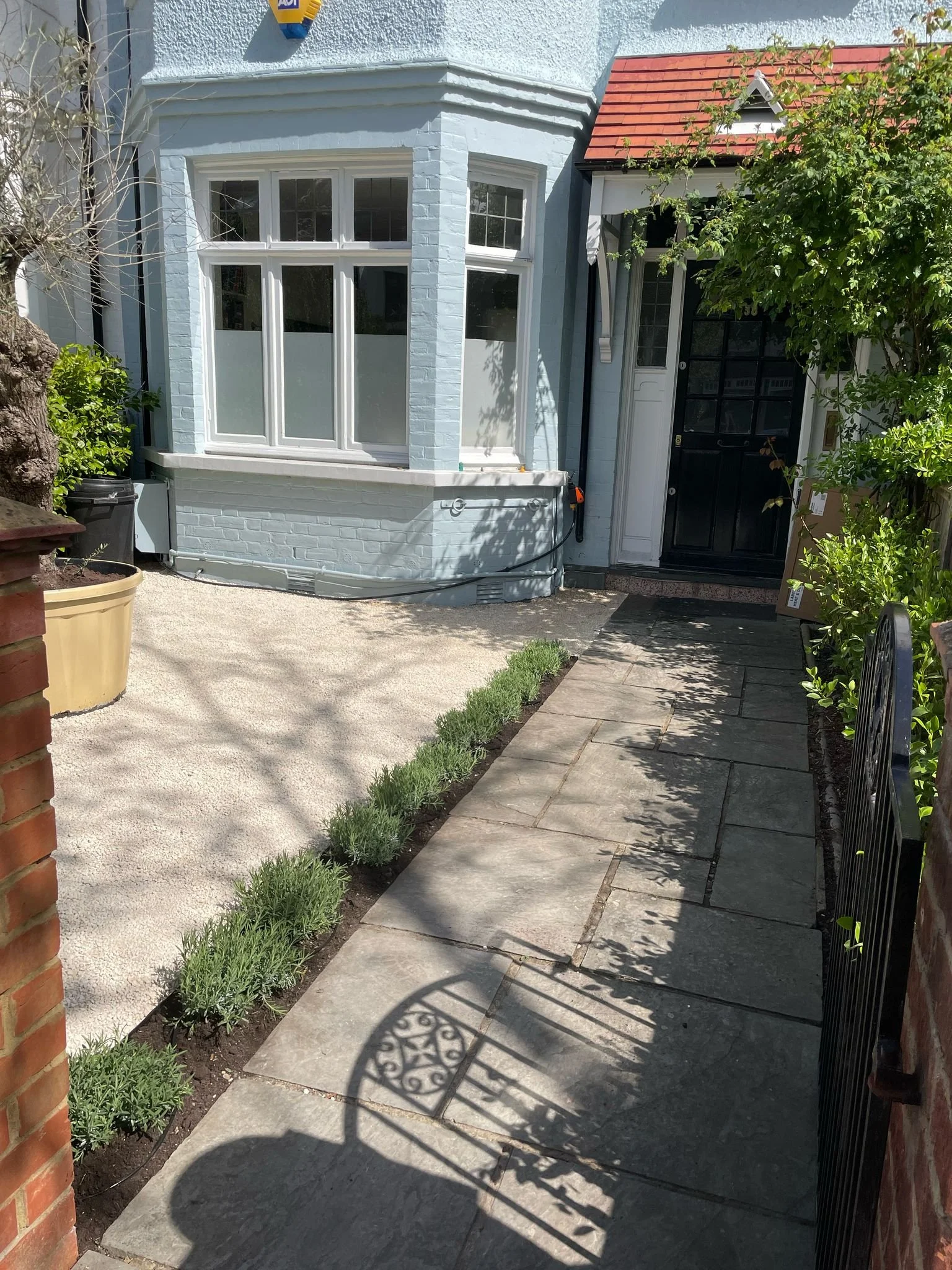 Front entrance of a light blue house with a bay window, a small porch with a black door, concrete and stone pathway, small hedges, a shadow of a decorative iron gate, and a tree with green leaves.