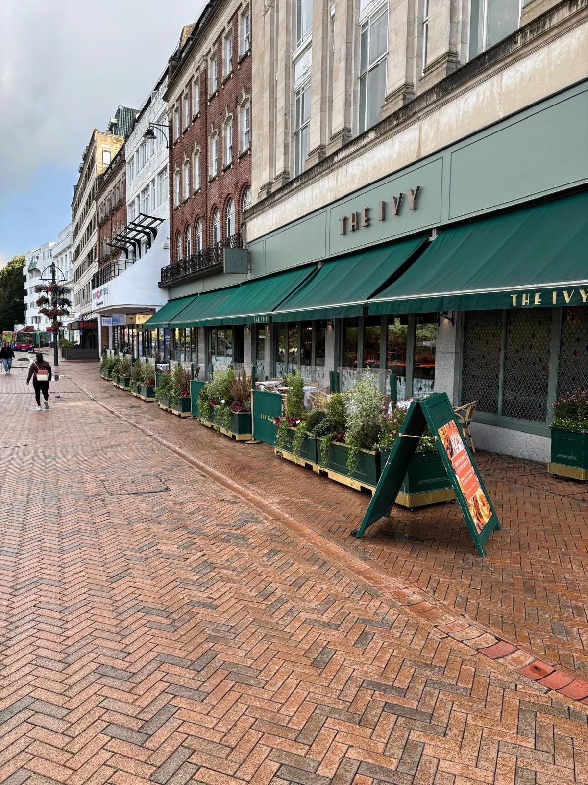 Street view of a restaurant named 'The Ivy' with outdoor seating and green awning, pedestrians walking on brick sidewalk.