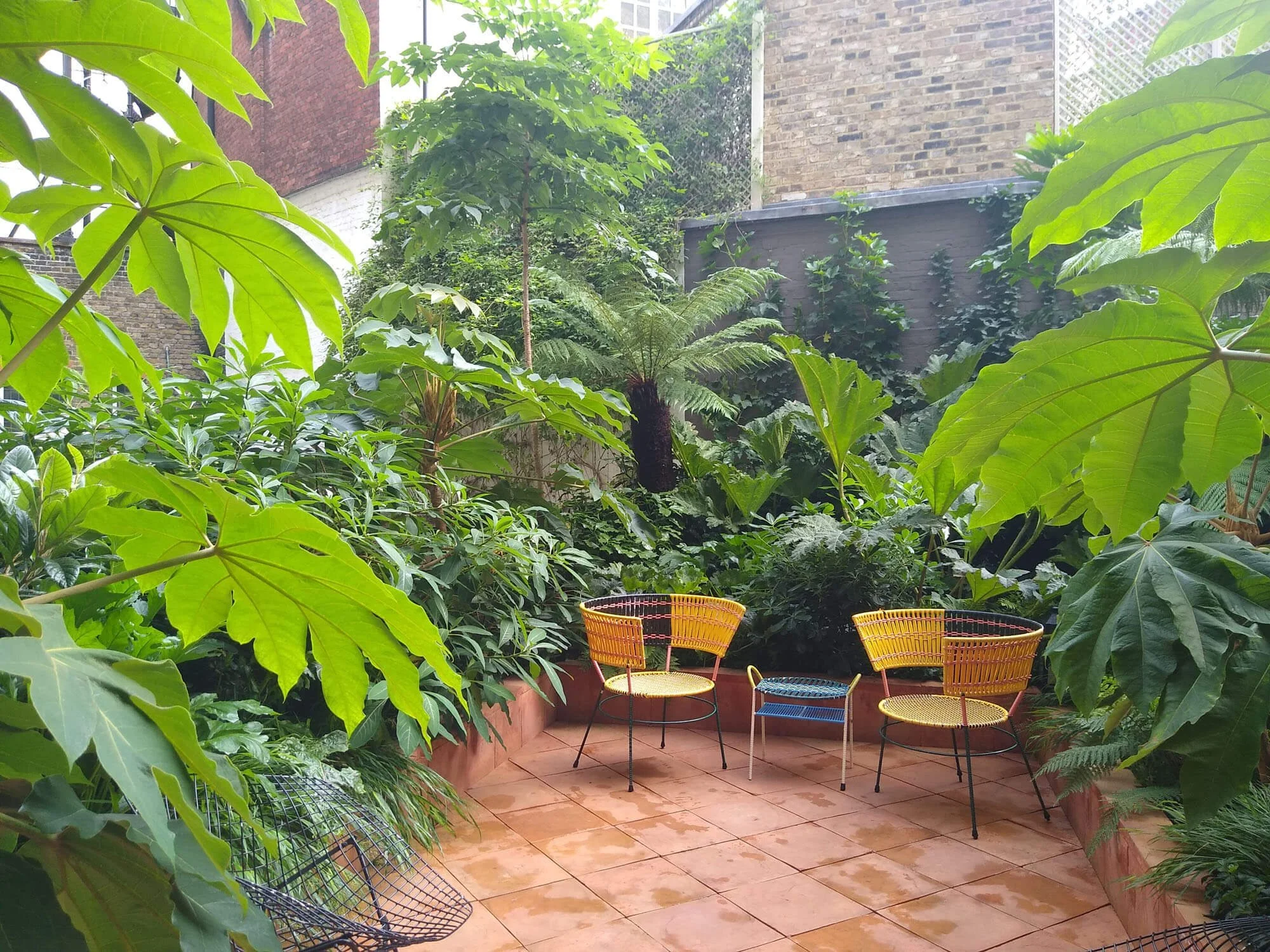 A small outdoor patio surrounded by lush green plants and trees, with colorful metal chairs and a brick floor.