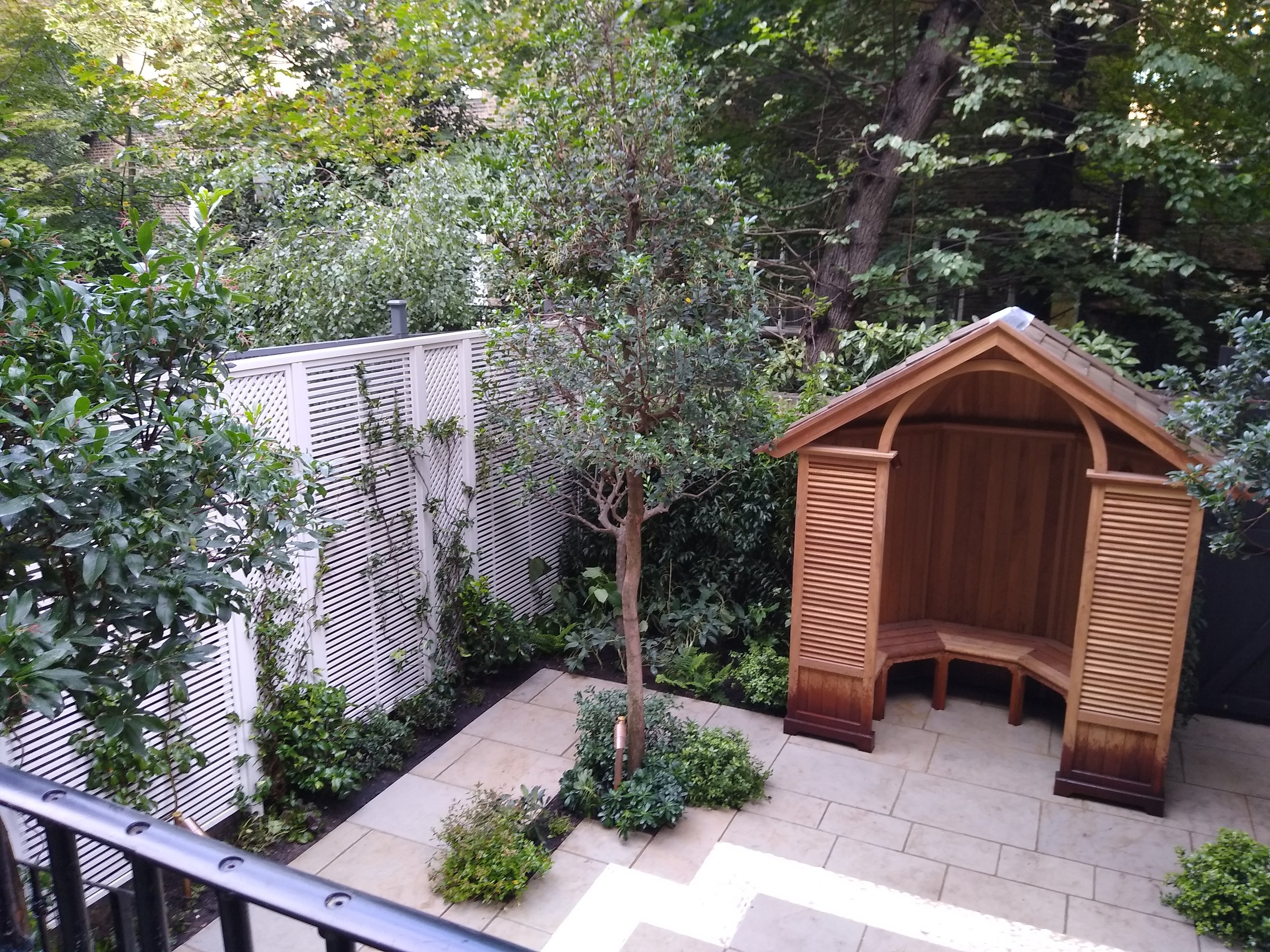 Small backyard with beige stone patio, lush green bushes, a small tree, a white fence, and a wooden shed with louvered doors.