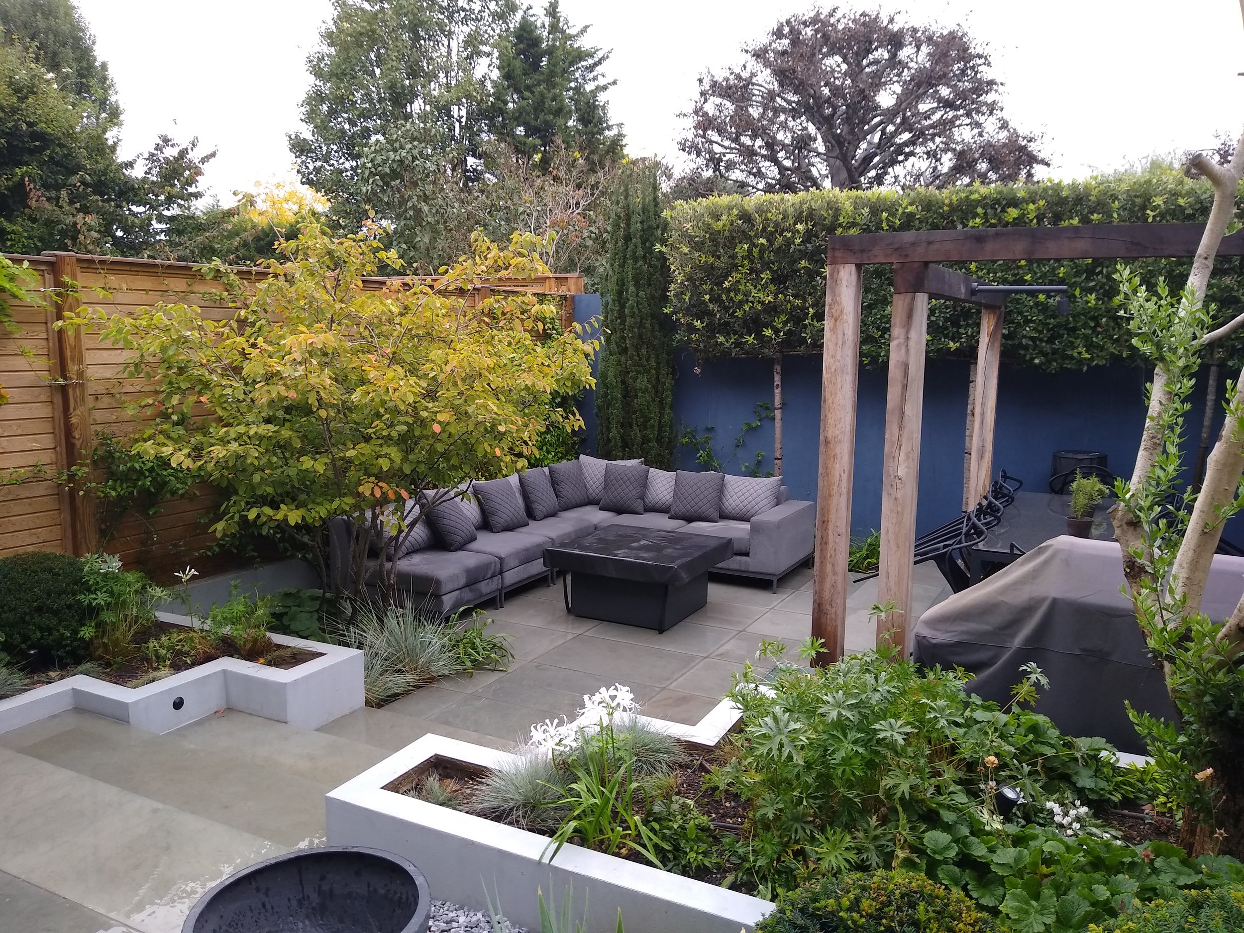 A cozy outdoor patio with a gray sectional sofa and matching pillows, a wooden pergola structure, potted plants, and a garden with various trees and shrubs, all enclosed by a wooden fence and a blue wall.