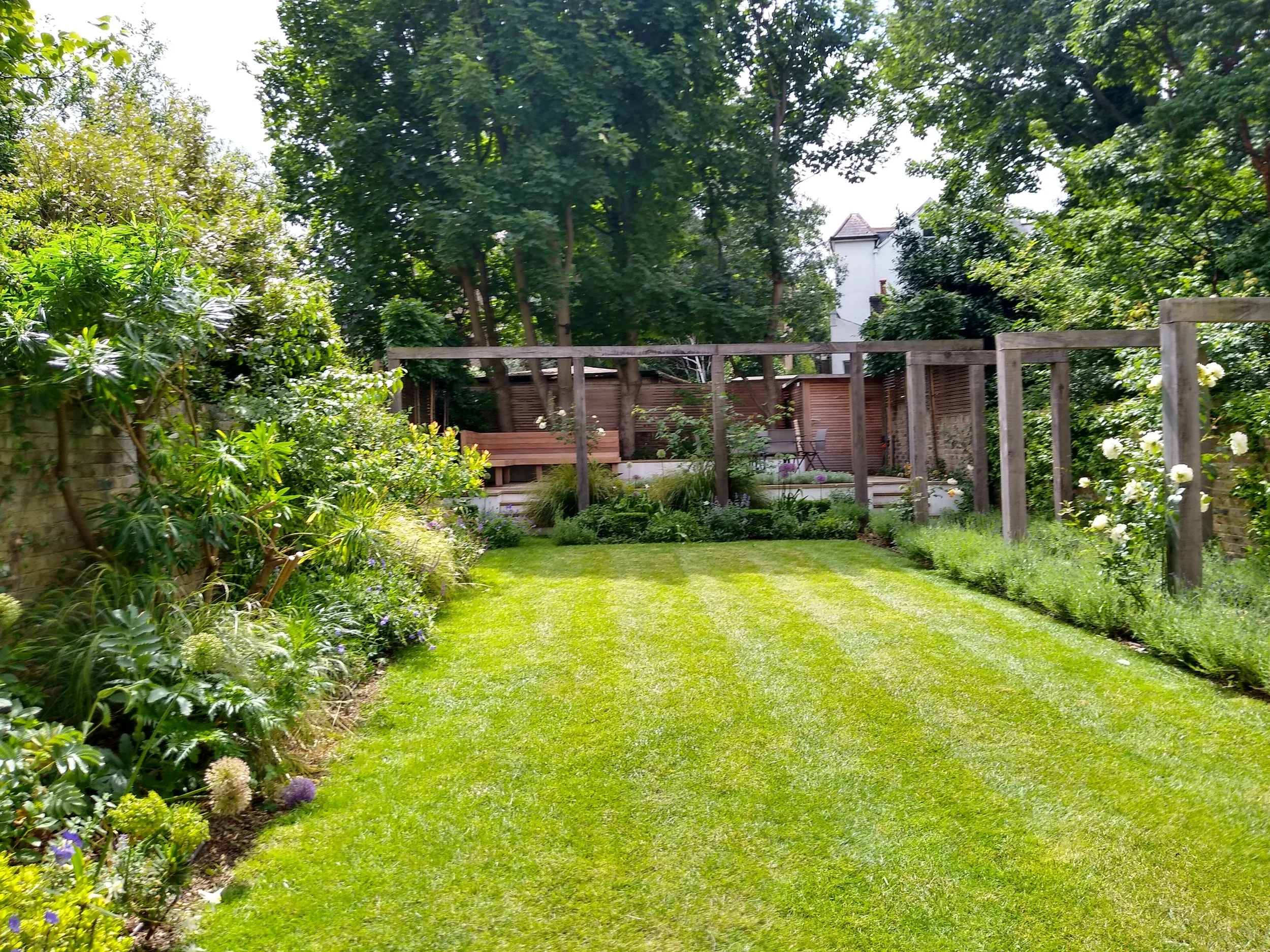 A well-maintained backyard garden with lush green grass, various plants along the sides, and trees providing shade. Wooden structures and a white house are visible in the background.