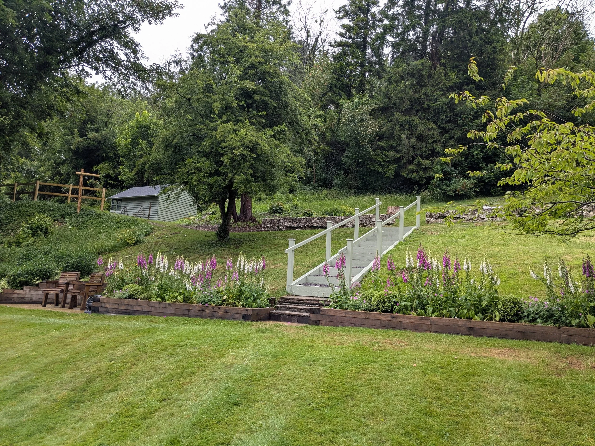 A backyard garden with a white wooden staircase leading up to a grassy area with a stone wall. The garden is bordered with purple and white flowers, and tall green trees surround the area. There are wooden benches on the left side and a small shed in