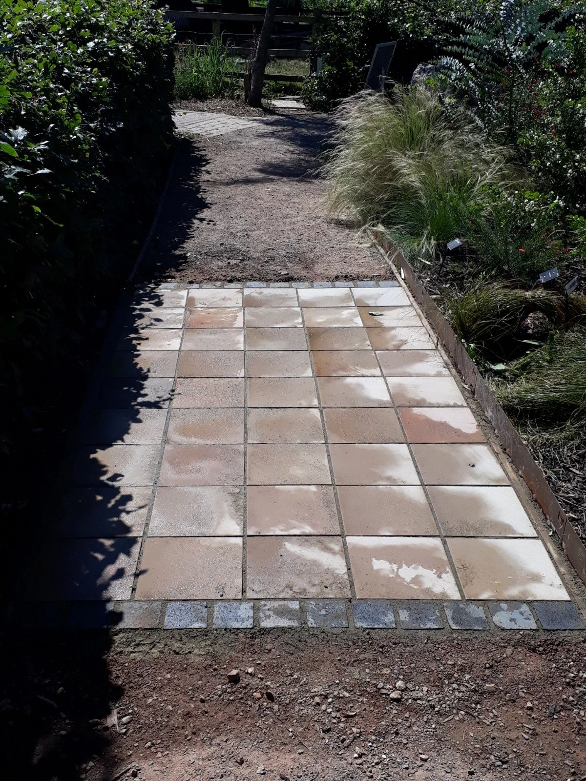 Patterned pathway being constructed with tiles, adjacent to a garden bed with ornamental grasses and plants, leading to a sidewalk with bare earth visible.