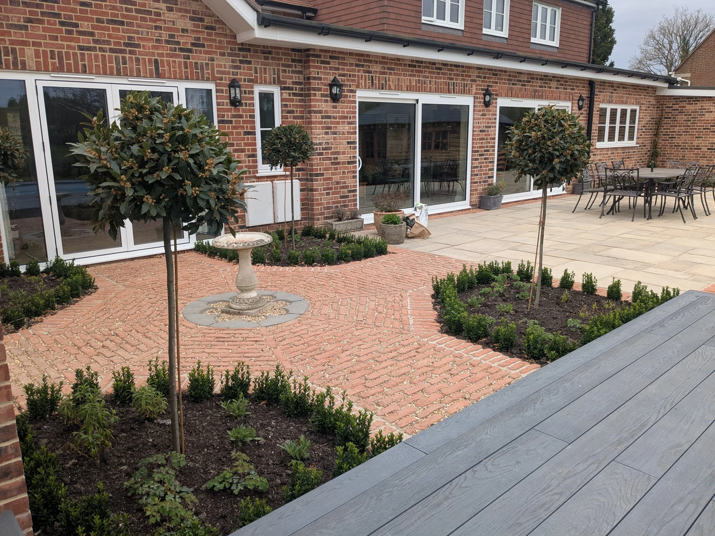 Backyard patio with brick and stone paving, two ornamental trees, various potted plants, garden bed with small shrubs, outdoor dining table with chairs, and sliding glass doors leading into a brick house.