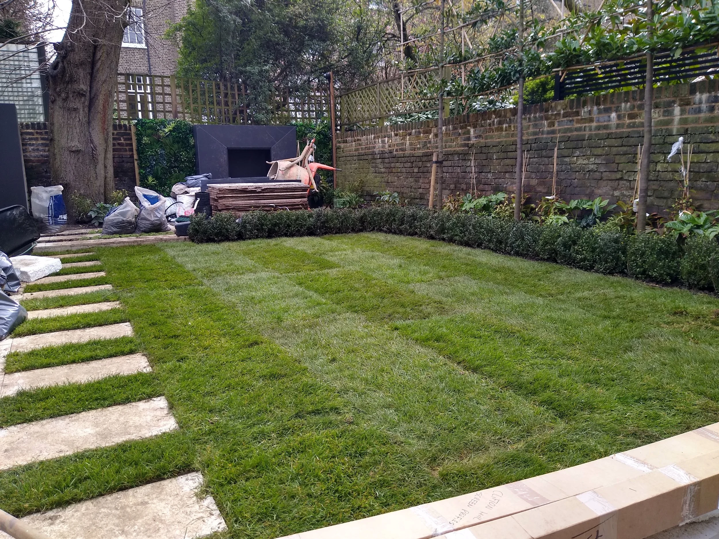 A backyard garden with a lawn, stone pathway, and gardening supplies, including bags and a shovel, near a brick wall and trees.