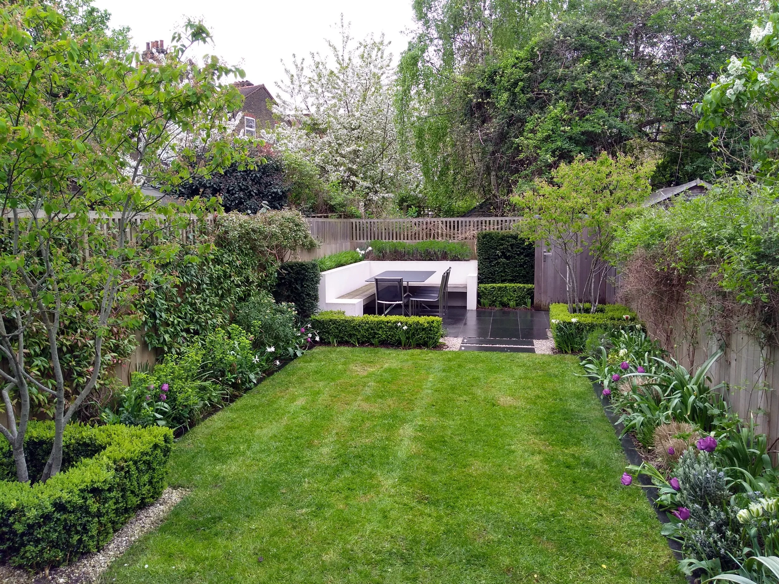 A well-maintained backyard garden with a green lawn, flower beds with purple and white flowers, small trees, and shrubs. There is outdoor seating with a black table and chairs on a tiled patio area, enclosed by a wooden fence, with a backdrop of leaf