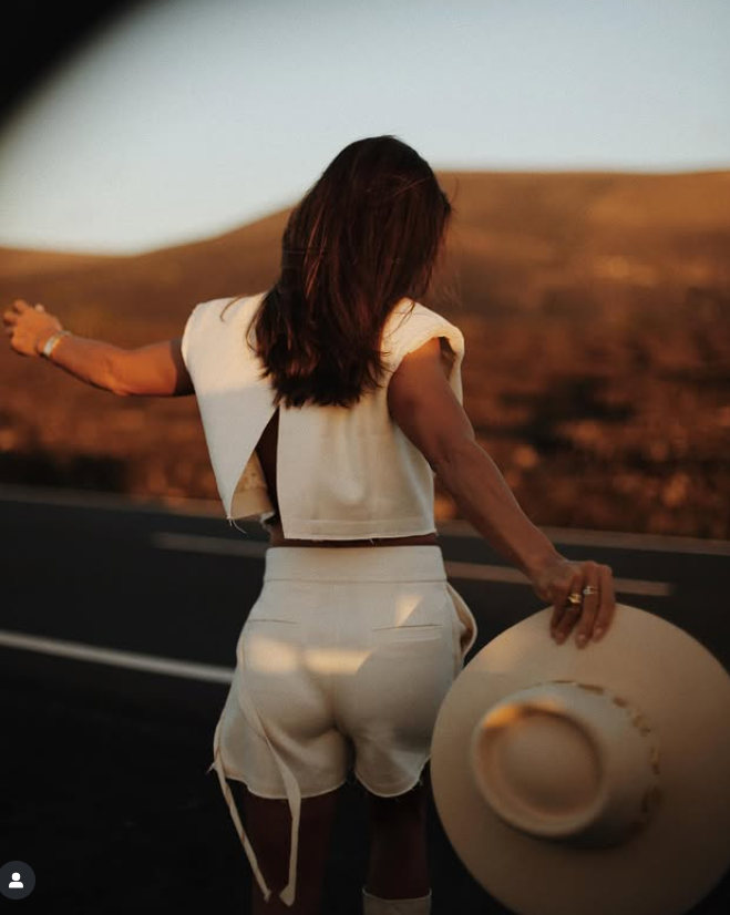 Woman standing on a road at sunset, holding a wide-brimmed hat, with her back to the camera.