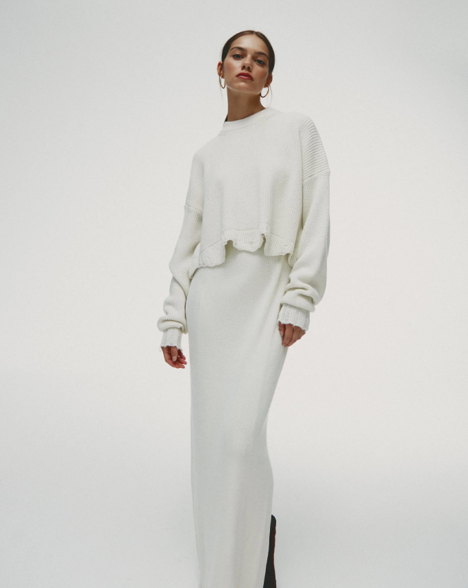 A woman wearing a white oversized knitted sweater and a long white skirt, standing against a plain white background.