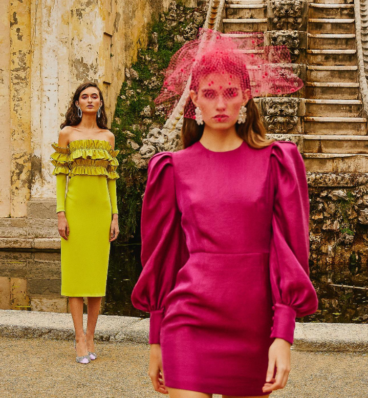 Two women in colorful dresses and vintage accessories stand outdoors in front of an old stone staircase and wall. One woman in the foreground wears a magenta dress with puffed sleeves and a pink veil, while the other in the background wears a lime green dress with ruffles.