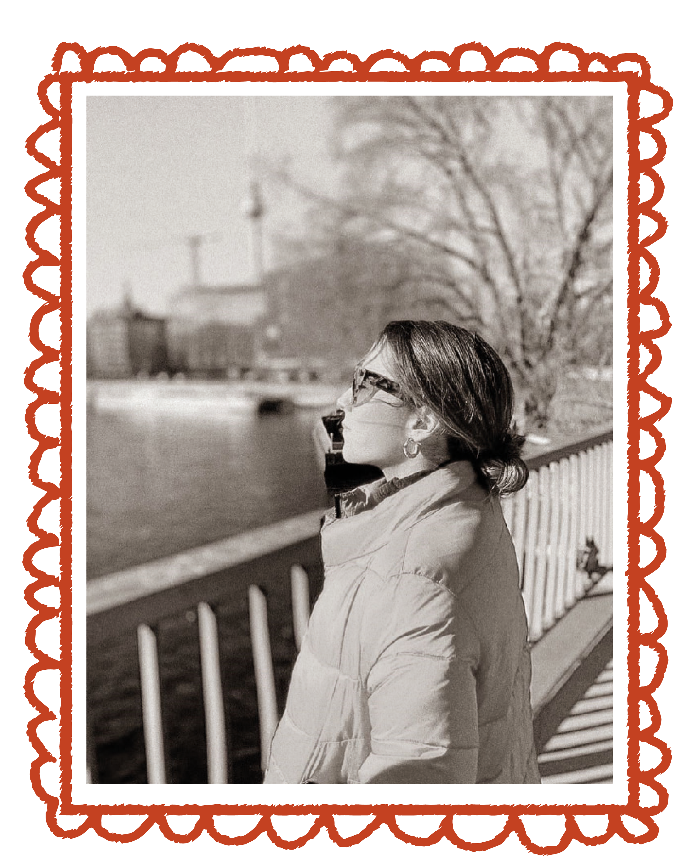 A woman wearing sunglasses and a coat, standing by a river on a sunny day, with leafless trees and buildings in the background.