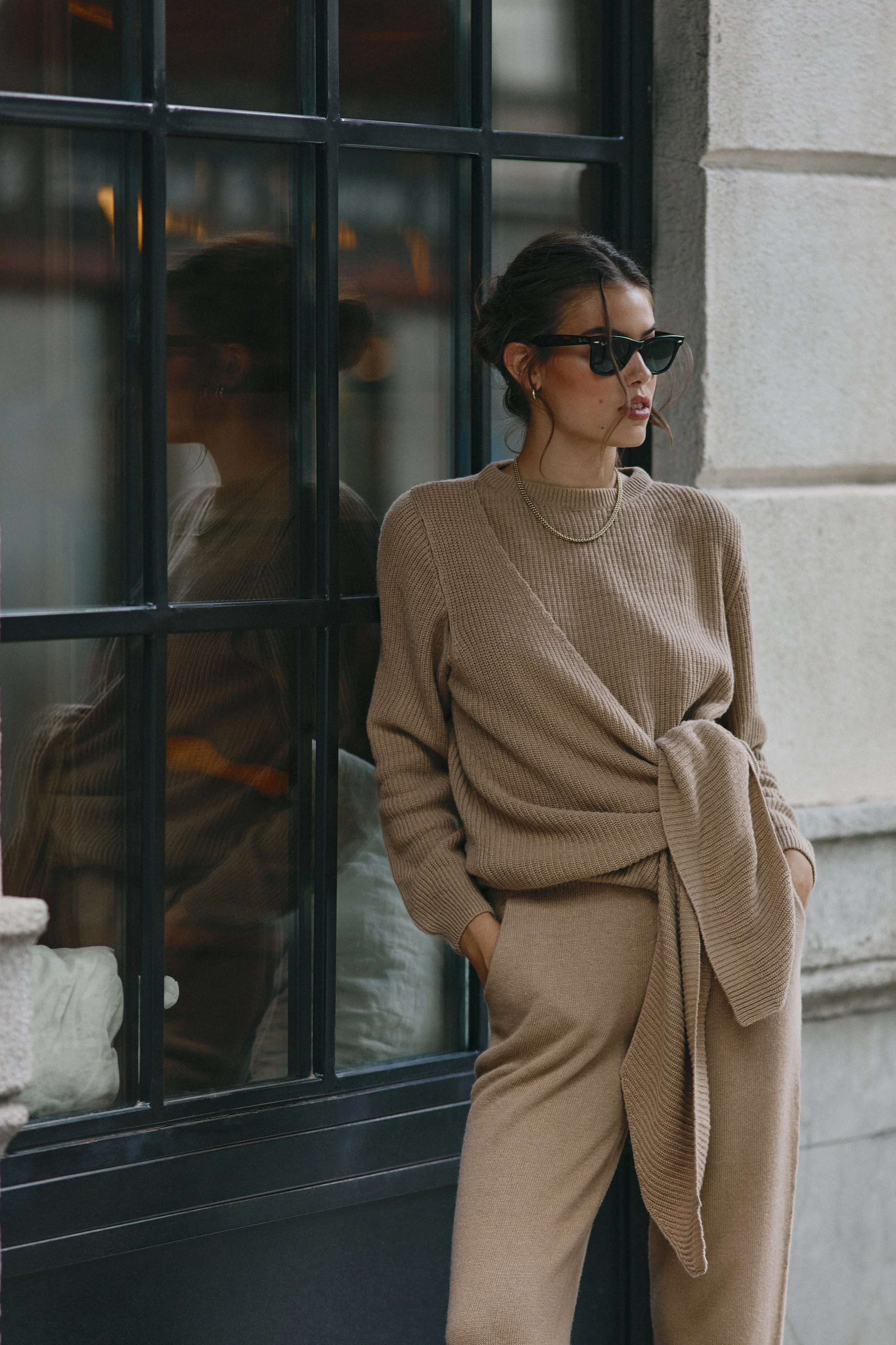 A woman with dark hair wearing sunglasses, a beige knit top with a knot at the waist, matching beige pants, and jewelry, standing outside a building with large black-framed glass windows and a light-colored stone wall.