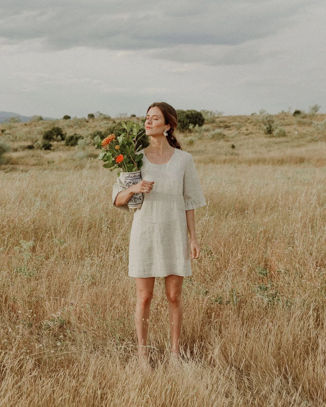 A woman in a beige dress standing in a field of tall grass, holding a bouquet of flowers, with a cloudy sky in the background.