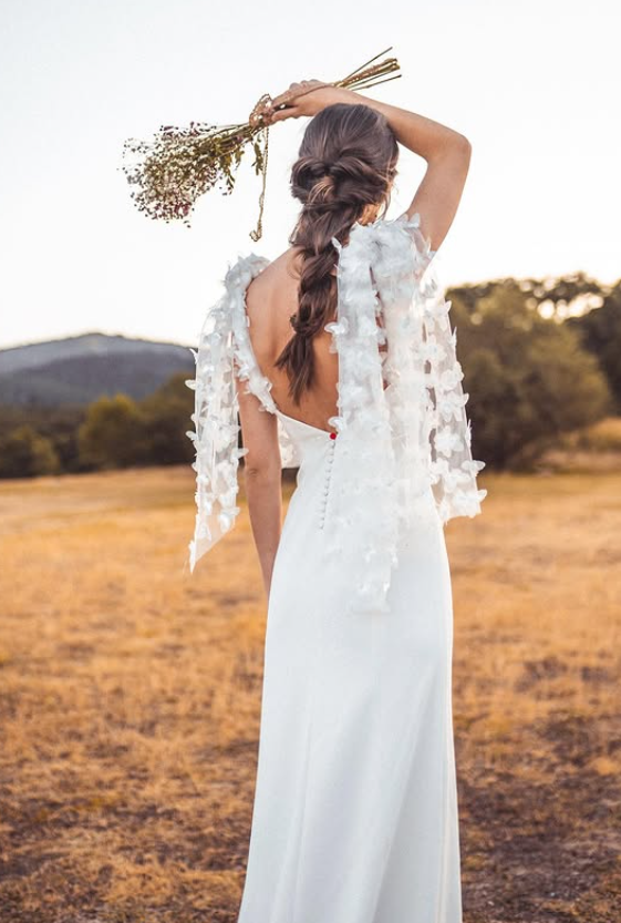 A woman in a white dress standing in an open field during sunset, holding a bouquet of flowers above her head, with her back facing the camera.