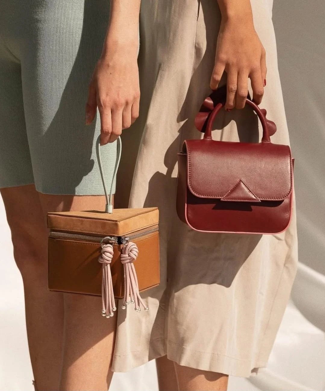Two women, one wearing a green skirt and the other beige pants, are holding handbags, one tan with a top handle and knotted strap, the other red with a top handle and a triangular detail on the flap.