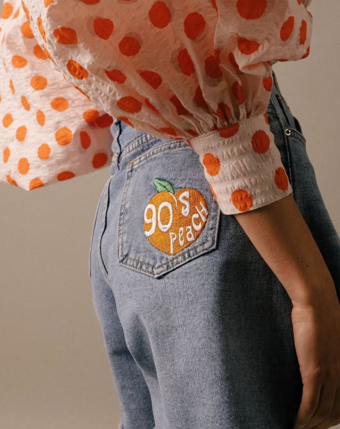 Close-up of a person's back wearing light blue jeans with a peach-shaped patch that reads "90s Peach" and a pink sleeveless top with orange polka dots.