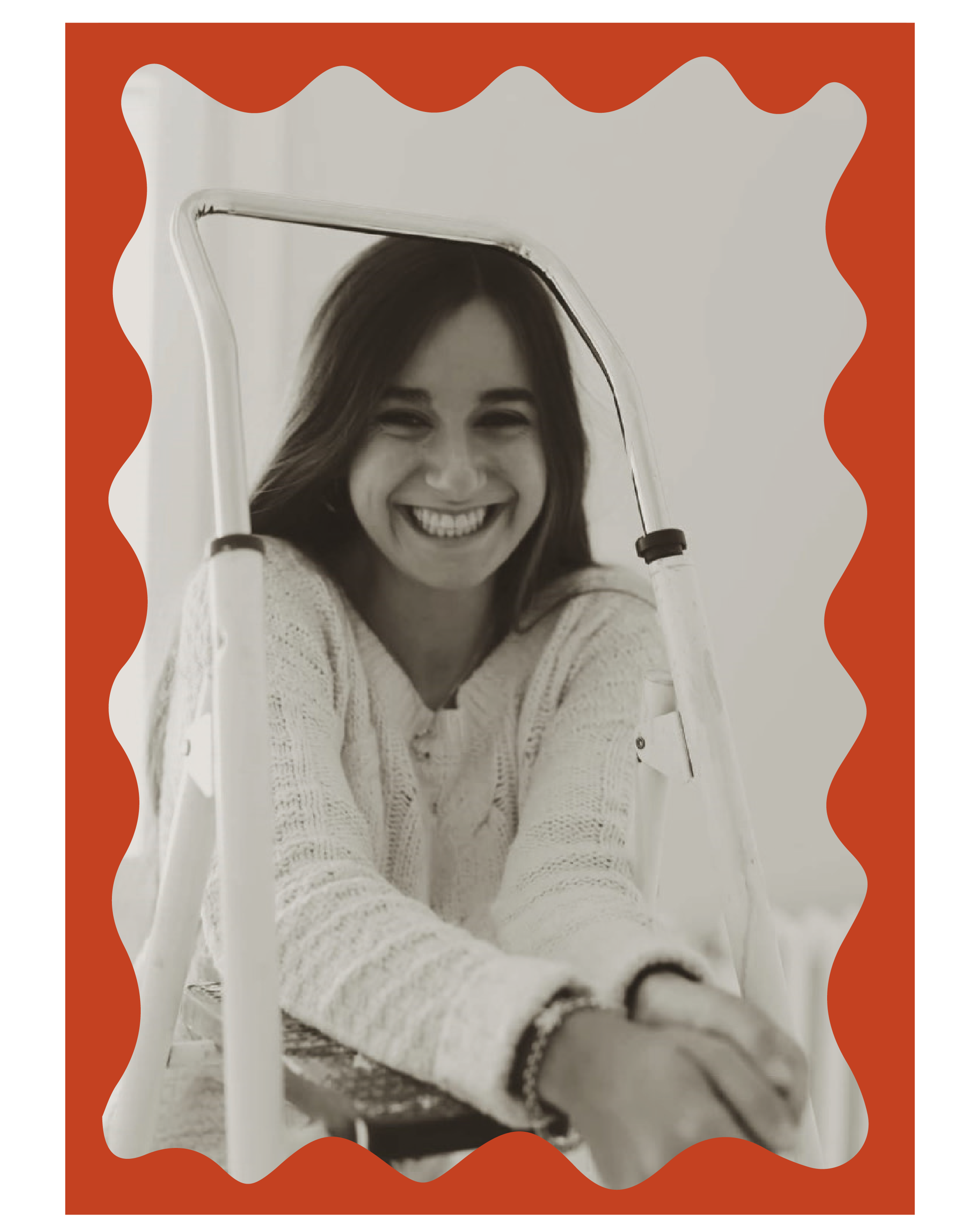 A young woman smiling through a mirror framed with a red wavy border, sitting on a chair, wearing a white knitted sweater.
