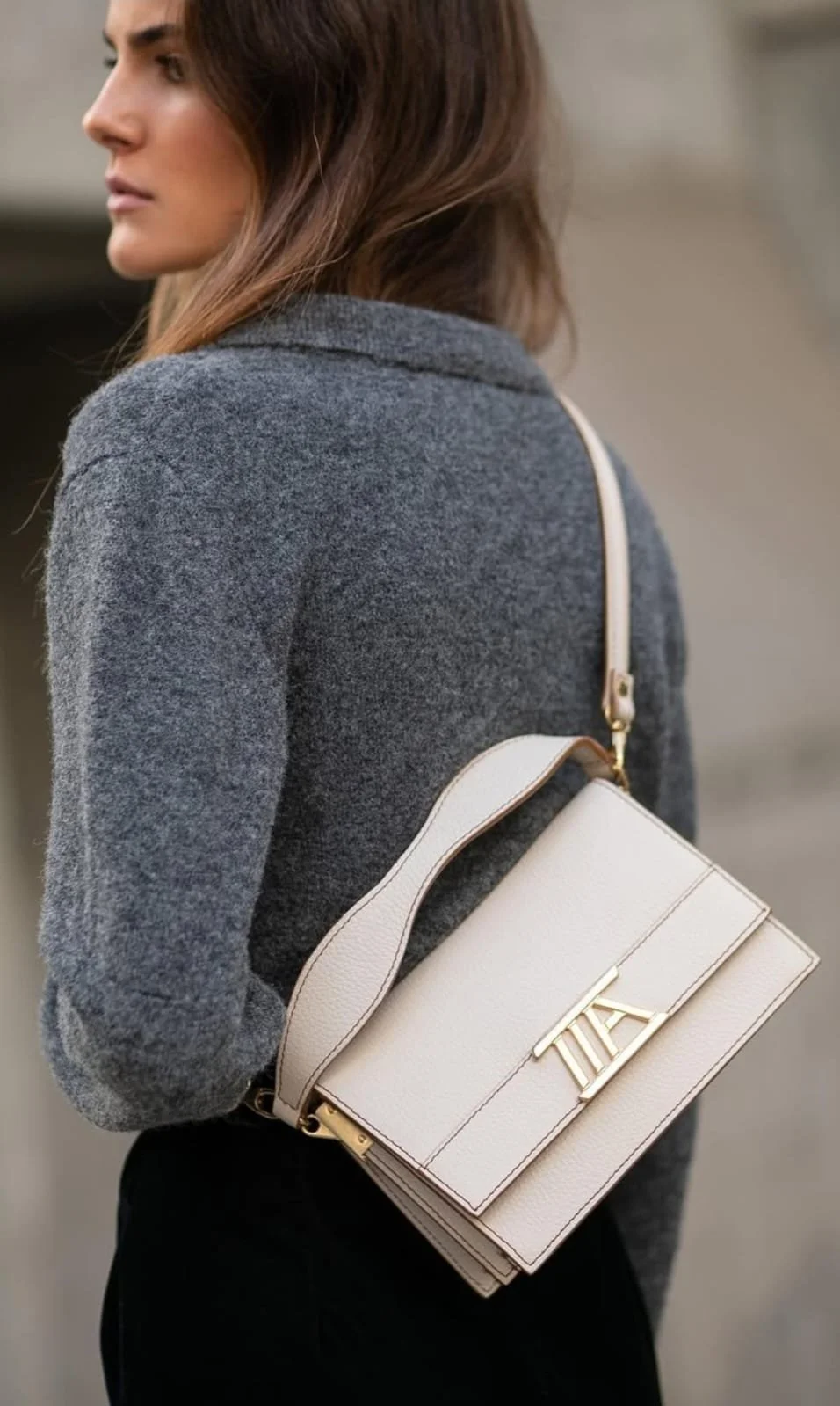 A woman with long brown hair wearing a gray jacket and carrying a white handbag with a gold logo on a beige strap.