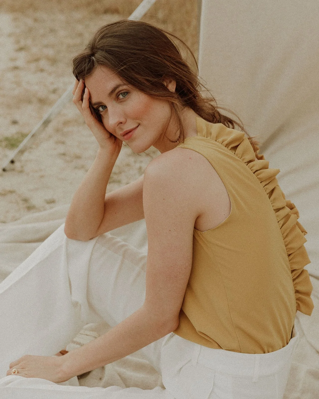 A woman with brown hair and blue eyes sitting on a white fabric in a sandy outdoor setting, wearing a yellow sleeveless top with ruffles on the back and white pants, looking at the camera with her hand on the side of her face.