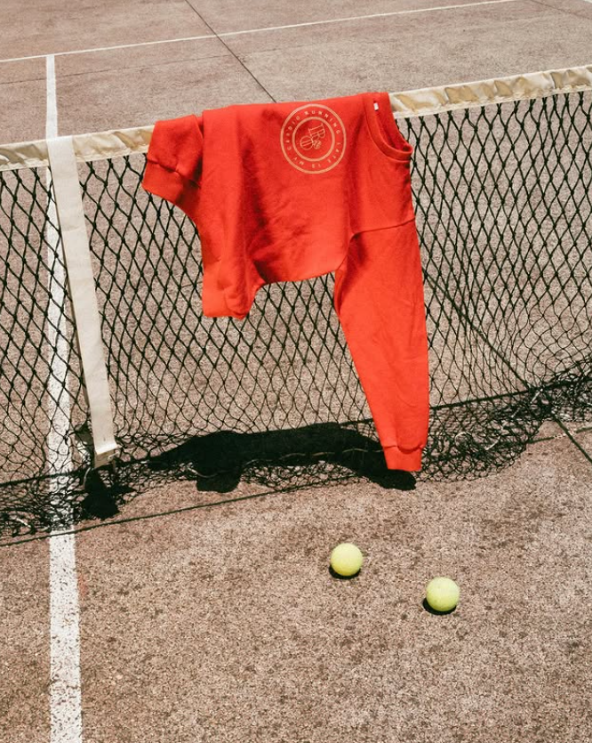 A red sweatshirt hanging on a tennis net with two tennis balls on the ground nearby.