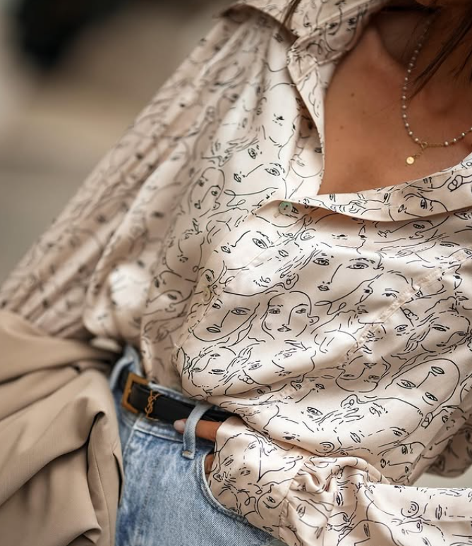 Close-up of a person wearing a cream-colored shirt with a pattern of abstract face sketches, paired with light blue denim jeans, a black belt with an orange logo, and a gold necklace. A beige jacket is draped over their arm.
