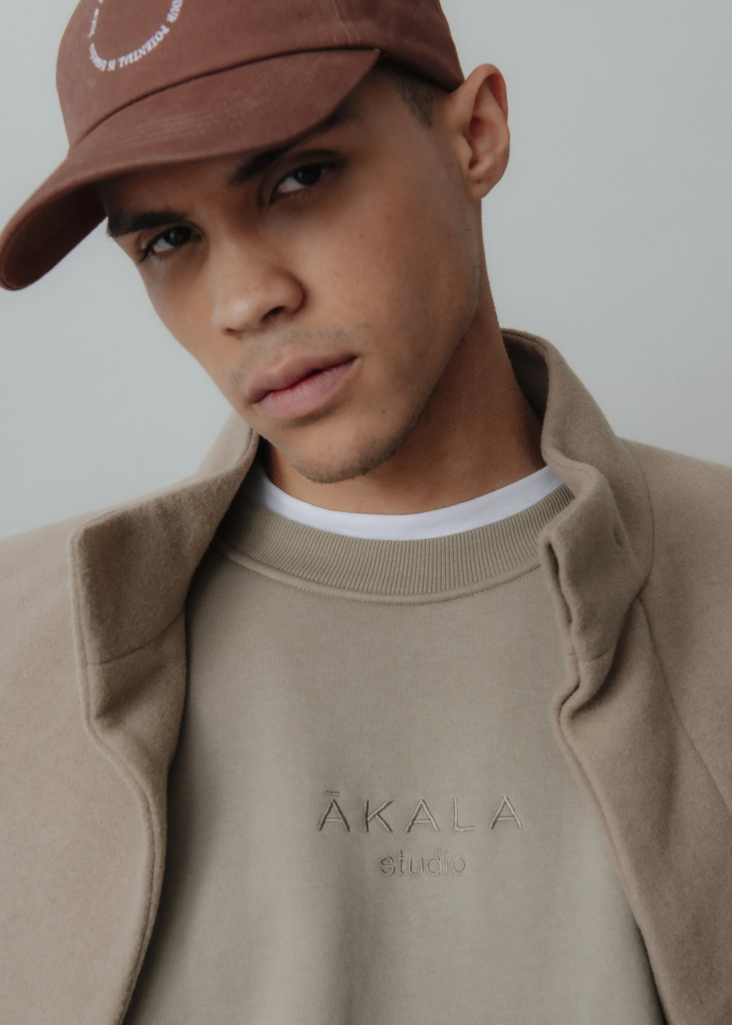 Close-up of a young man wearing a brown baseball cap, beige coat, and a beige sweatshirt with the words 'ÁKALA studio' embroidered on it, standing against a plain white wall.