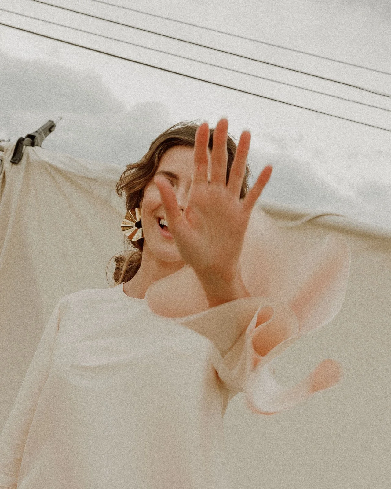 A woman smiling with a large earring, reaching out with her hand towards the camera, in an outdoor setting with a neutral background and overhead wires.