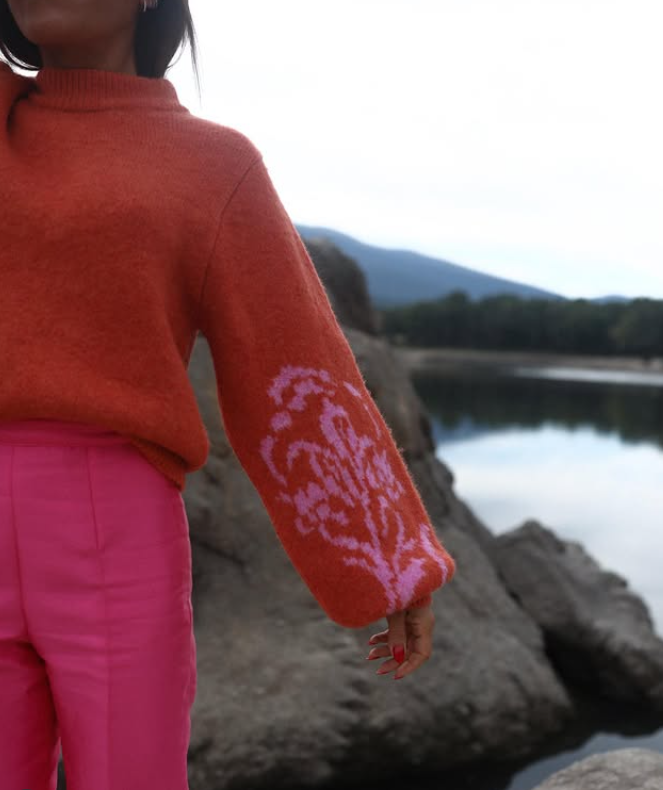 Person wearing an orange sweater with a pink design on the sleeve and pink pants, standing near a body of water with rocks and mountains in the background.