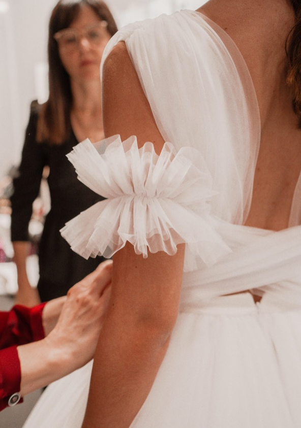 A woman wearing a white dress with ruffled sleeves is being assisted with fitting in a fitting room. In the background, another woman with glasses is visible.