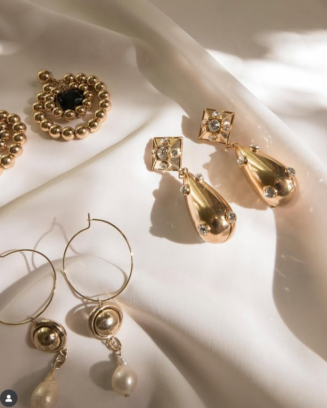 Close-up of gold and pearl earrings and gold jewelry, arranged on white fabric with shadows and lighting.