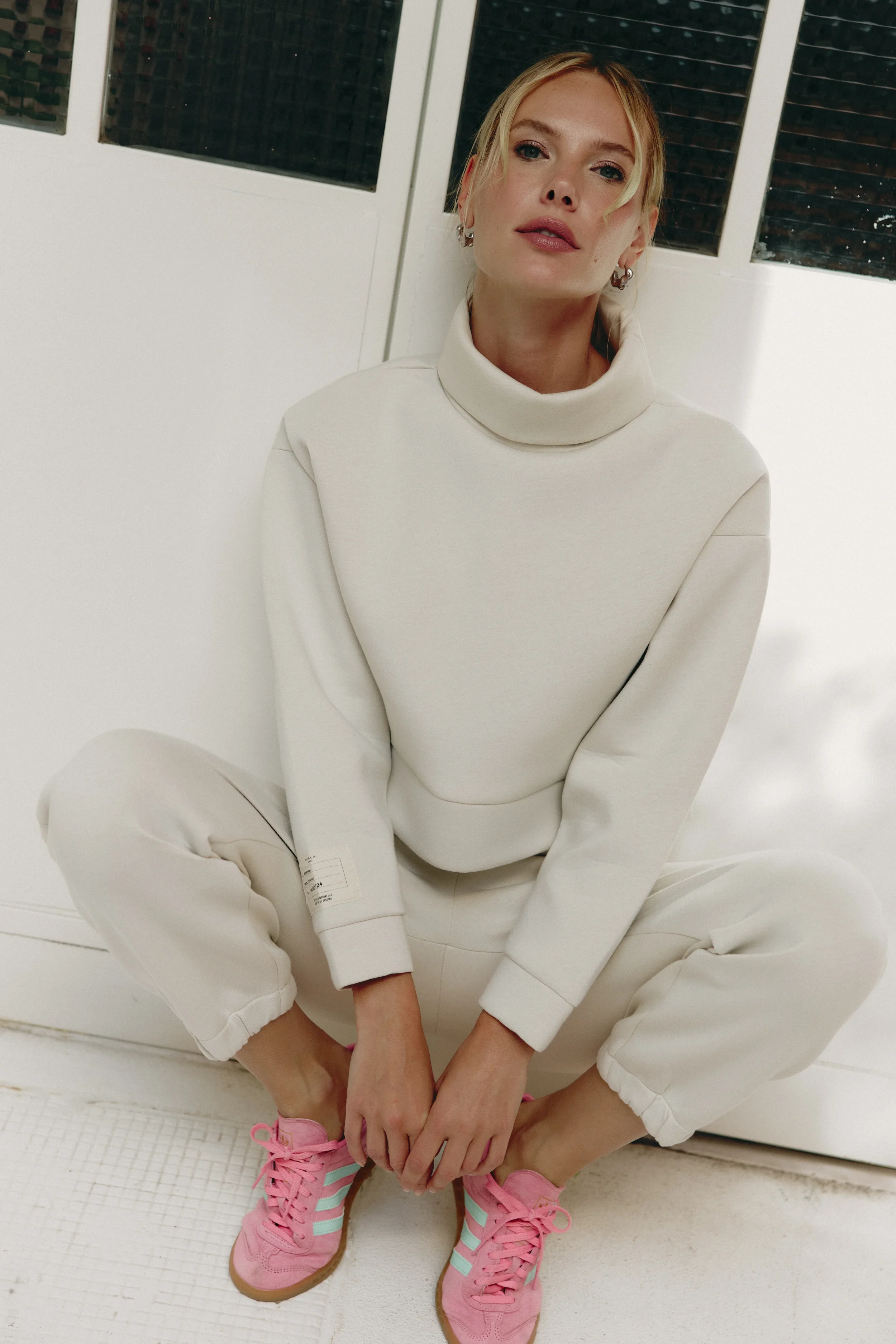 A woman with blonde hair tied back, wearing a white turtleneck sweatshirt, matching white sweatpants, pink sneakers with blue stripes, sitting cross-legged on the floor in front of a white door with windows.