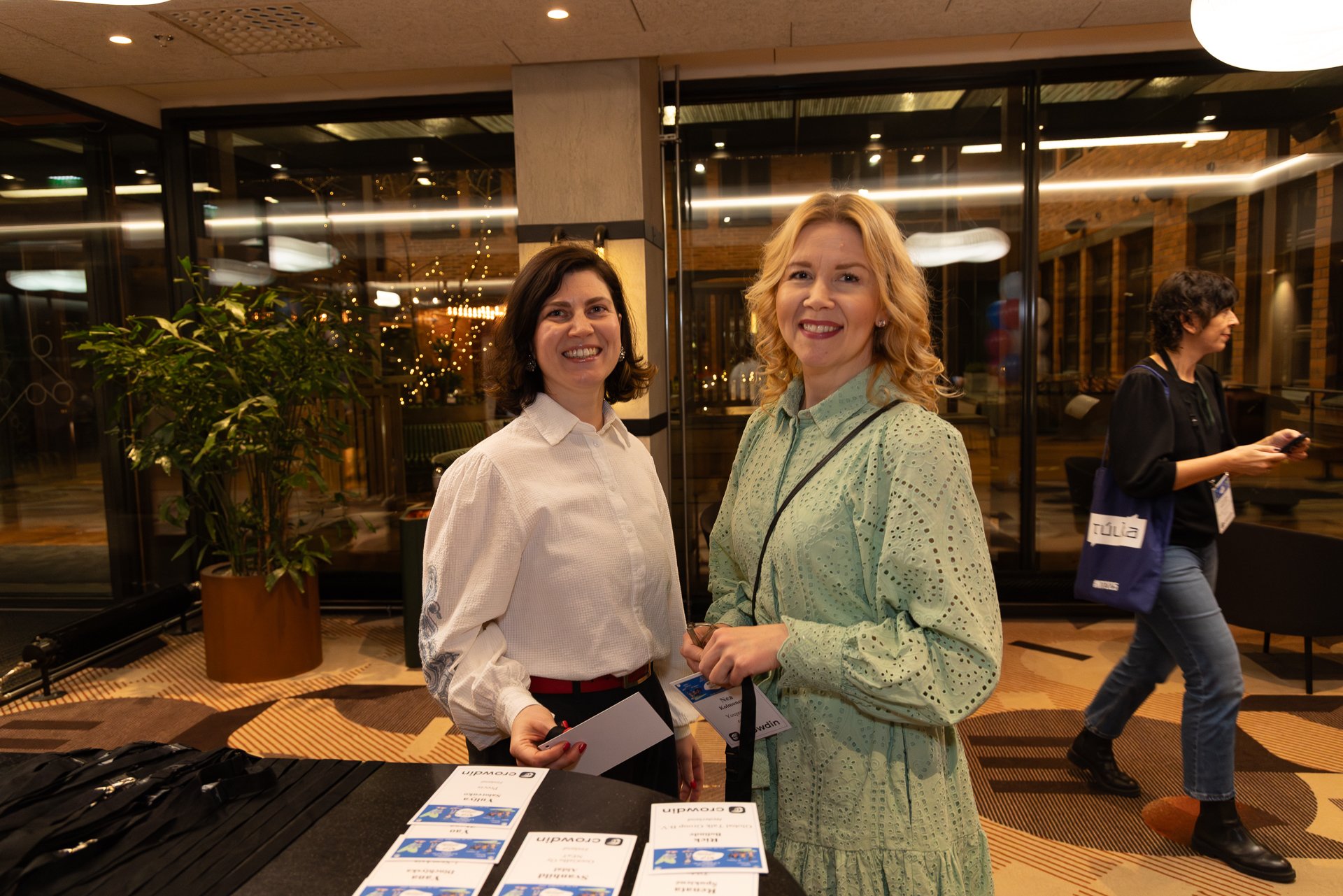 Kaksi naista hymyilevät ja poseeraavat sisätiloissa, lehtisiä pöydällä, muita ihmisiä taustalla kävelemässä.