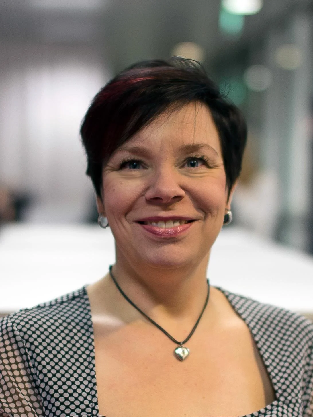 Hän on hyvin pukeutunut ja hymyilevä nainen, jolla on lyhyet tummat hiukset, ketjuhalaotsikoru ja musta-valkoinen pusero, taustalla hämärä sisätilo.