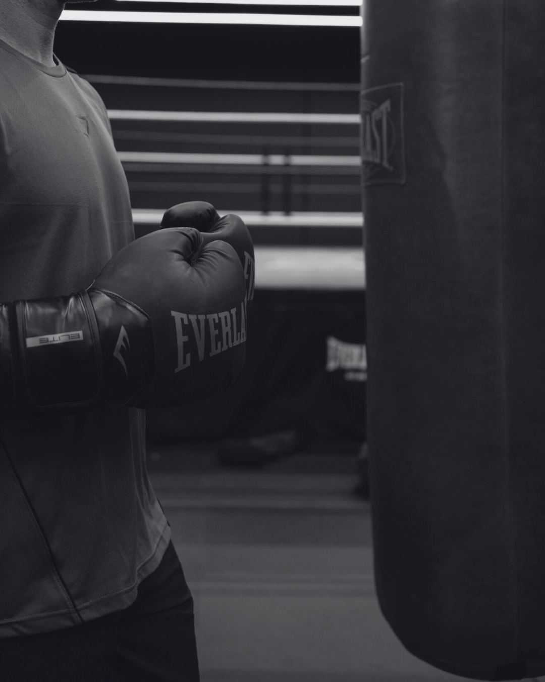 A boxer wearing black Everlast gloves and a sports shirt, standing near a punching bag in a gym, with a wall and exercise equipment in the background.