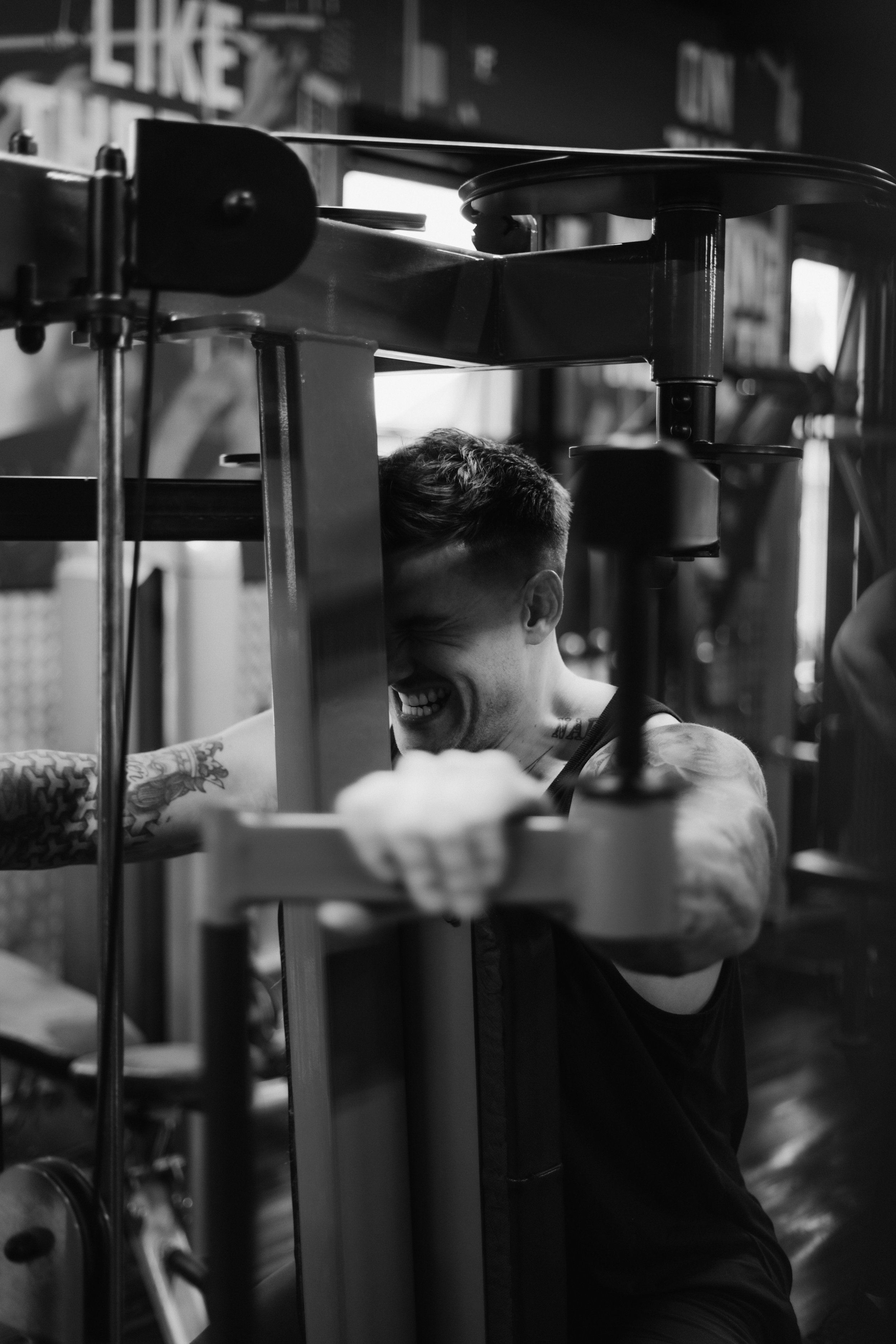 A joyful man with tattoos smiling as he leans against gym equipment in a fitness center.