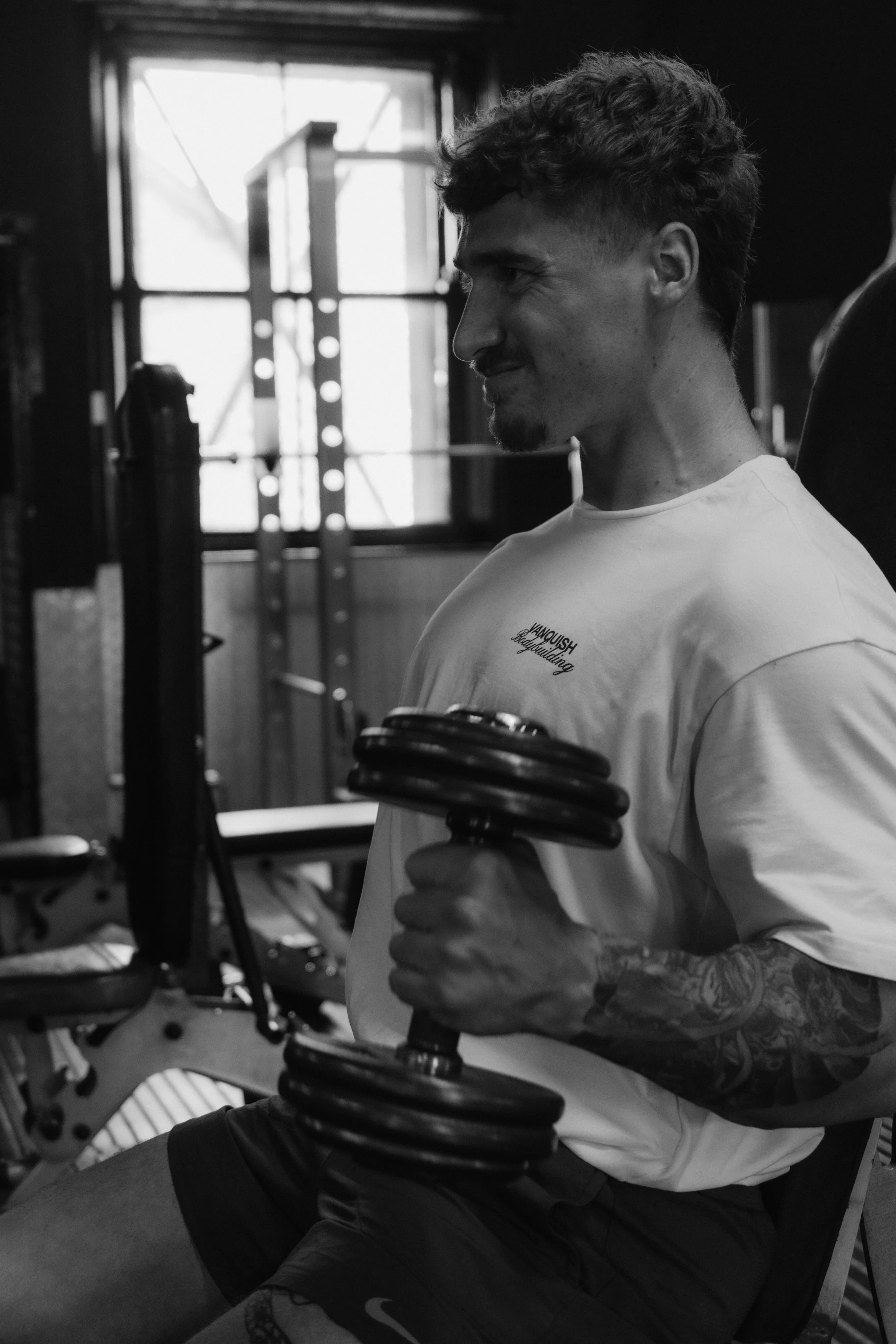 A man with curly hair and tattoos on his arm sitting in a gym, lifting a dumbbell while smiling.