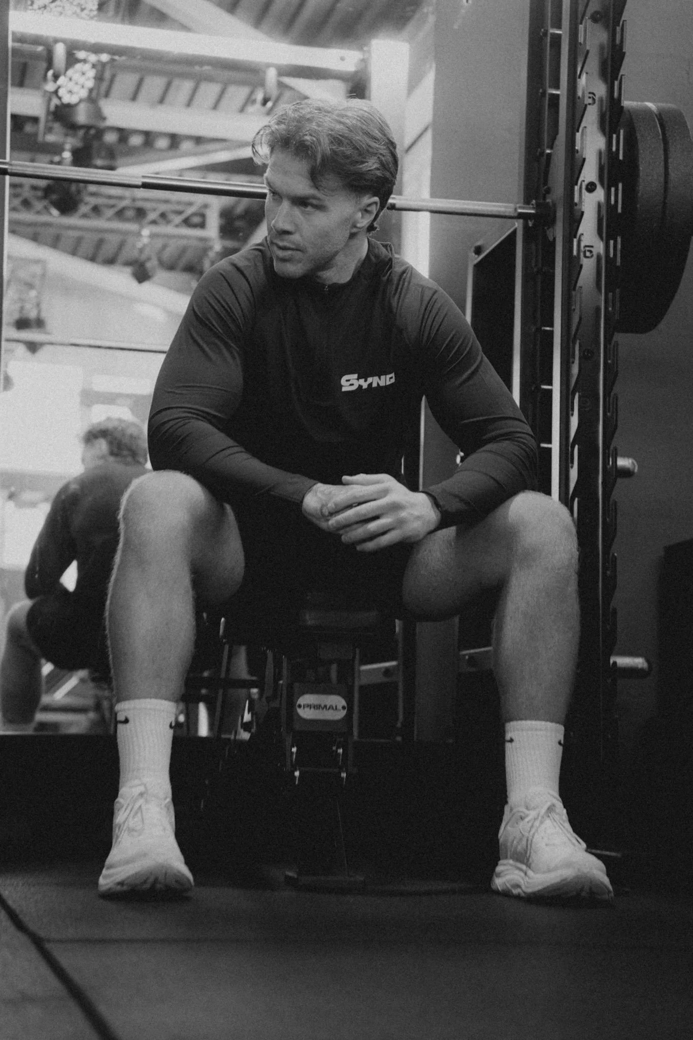 Young man sitting on a bench in a gym, wearing a long-sleeve shirt, shorts, and sneakers, with weightlifting equipment in the background.
