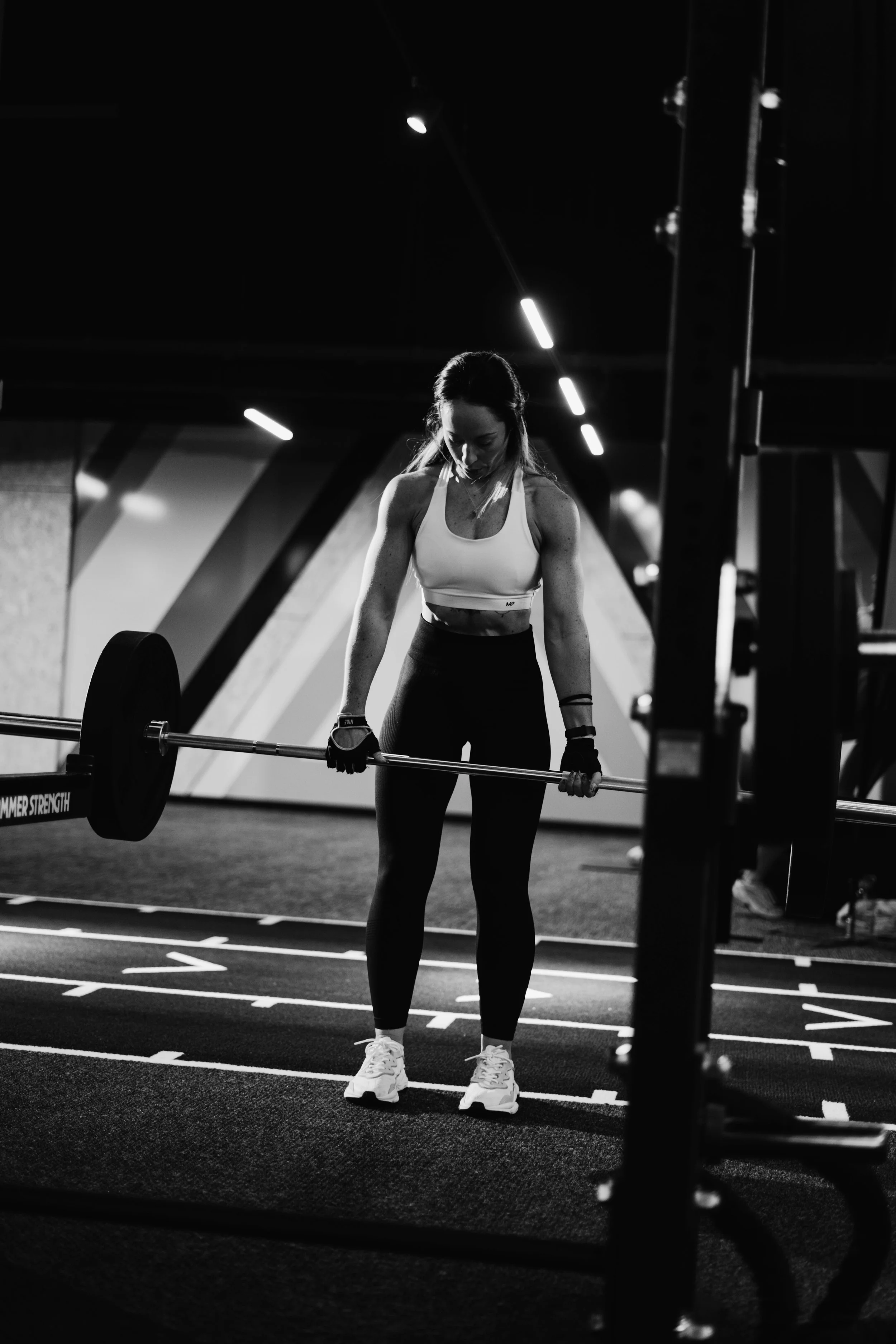 A woman lifting a barbell in a gym with a dark background.