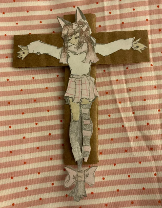 A paper craft drawing of a cartoon girl with animal ears, wearing shorts and standing on a bow-tie, mounted on a wooden cross, placed on a pink and white striped tablecloth with red polka dots.